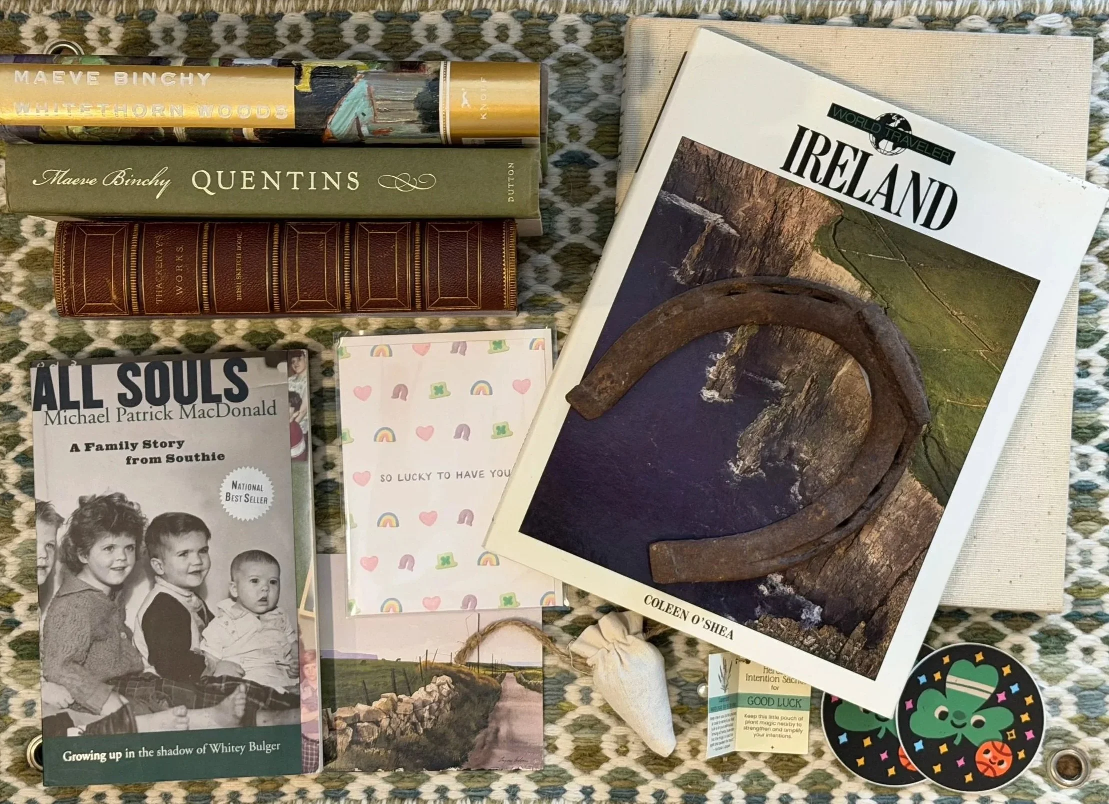 A collection of books on Ireland, family stories, and poetry, a postcard with rainbows and hearts, a horseshoe, a small stone, a tea bag, two round stickers with a green frog and Halloween pumpkins, and various decorative items on a woven cloth.