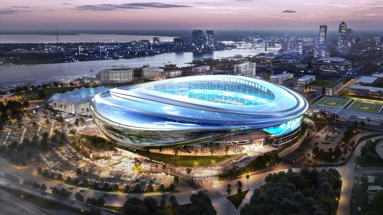 Aerial view of a futuristic football stadium illuminated at dusk in a cityscape with water and high-rise buildings in the background.