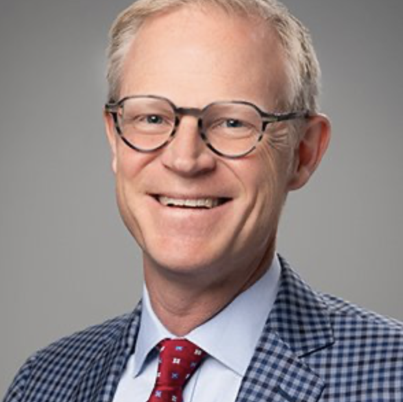 A middle-aged man with gray hair and glasses, wearing a checkered suit jacket, blue shirt, and red tie, smiling in front of a gray background.