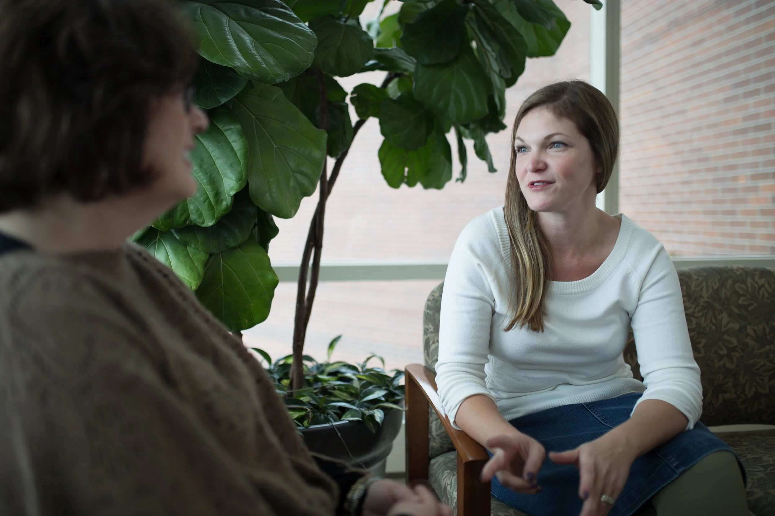 Two women sitting and talking in a room with large potted plants and a window showing a brick wall outside.