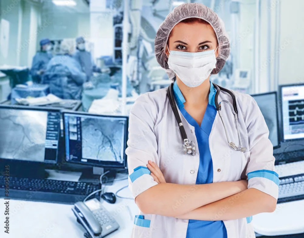 A female healthcare professional in a surgical mask and scrubs standing in a hospital control room with monitors and medical staff working in the background.