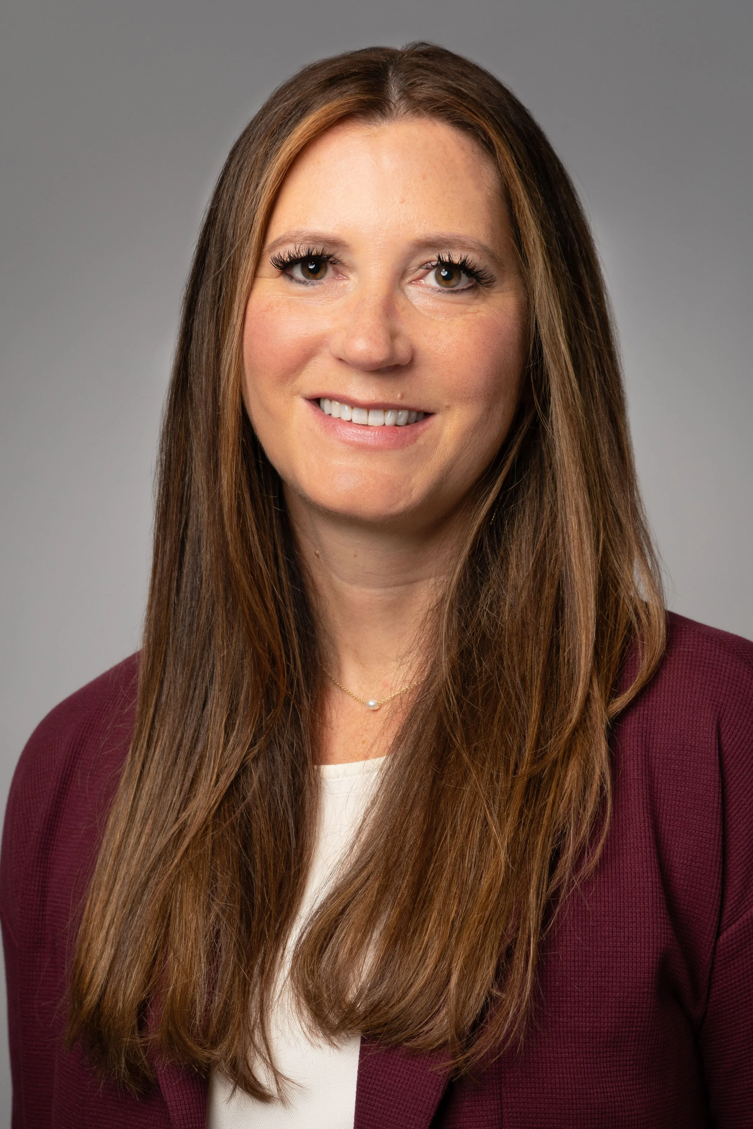 Professional headshot of a woman with long brown hair, wearing a burgundy blazer and white top, smiling, against a plain gray background.