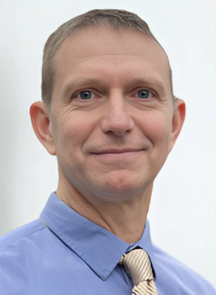 A professional headshot of a middle-aged man with short, light brown hair and blue eyes, wearing a blue dress shirt and a beige striped tie, against a plain light background.