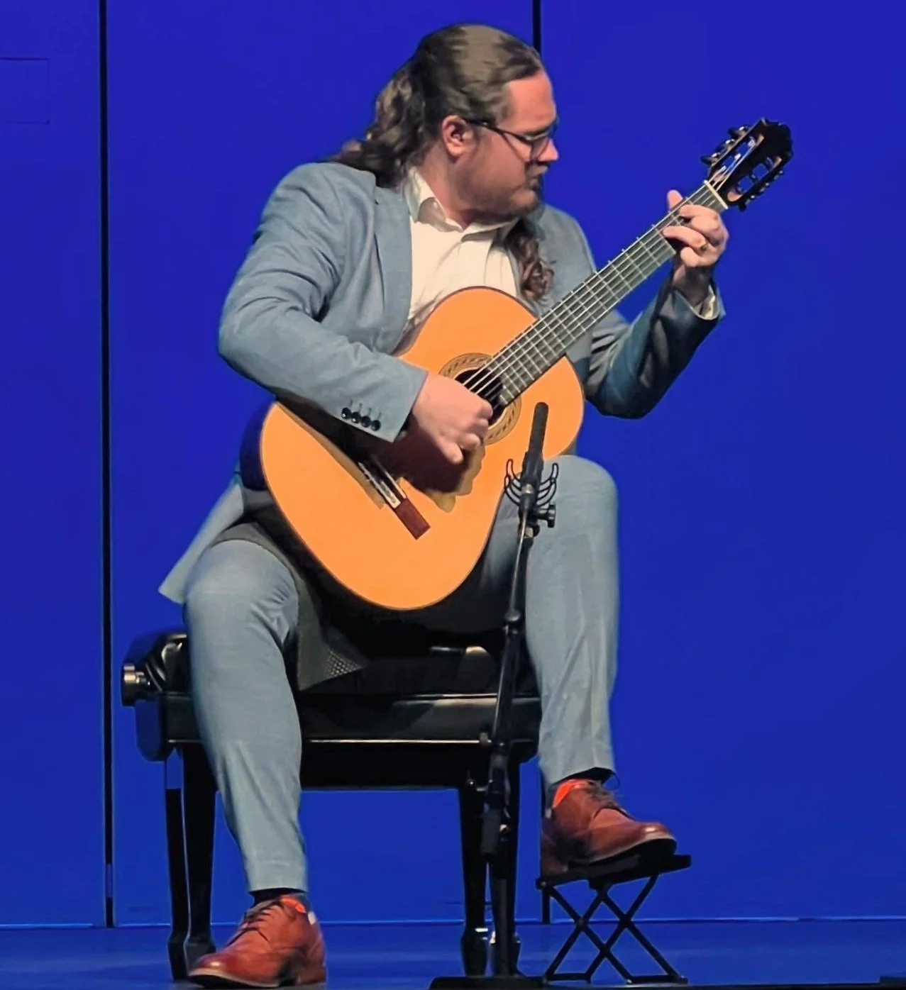 A man with long hair, glasses, and a grey suit jacket playing an acoustic guitar on a stage with a blue background.