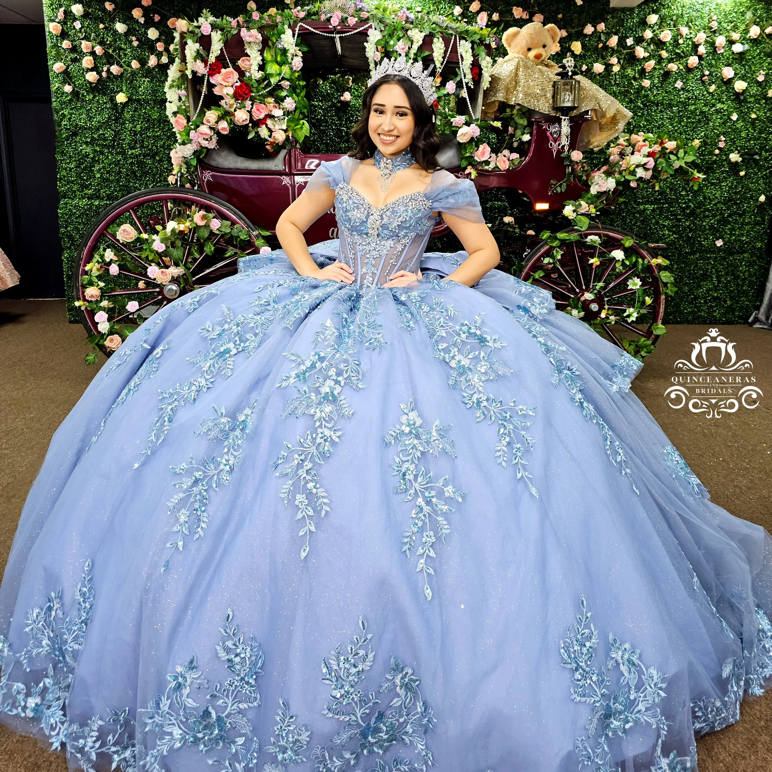 Young woman in a blue ball gown with floral embroidery, wearing a tiara, standing in front of a flower-decorated carriage and smiling.