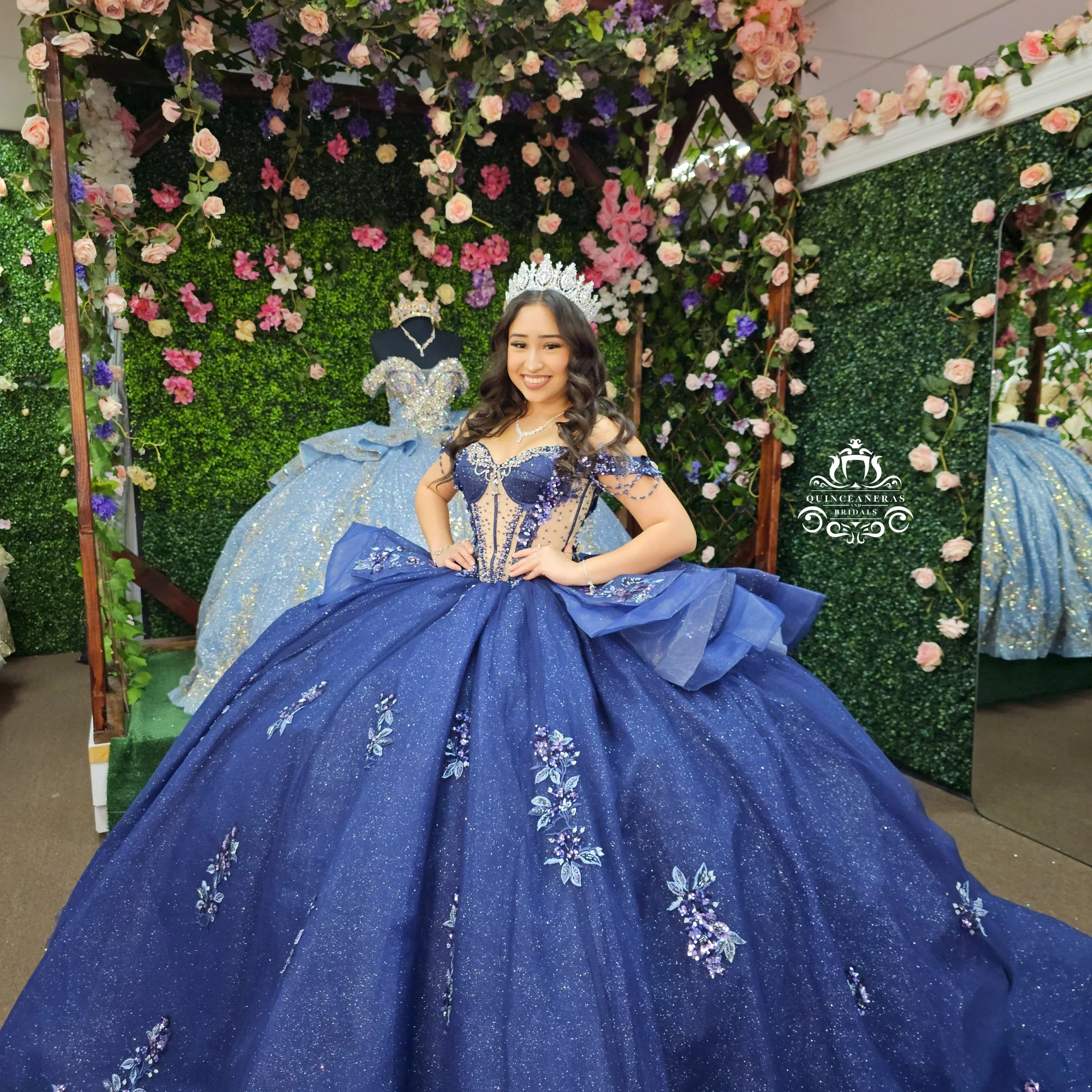 A young woman in an elegant, dark blue quince gown with floral embroidery and sparkles, wearing a tiara, in front of floral and greenery backdrop, smiling with hands on hips.