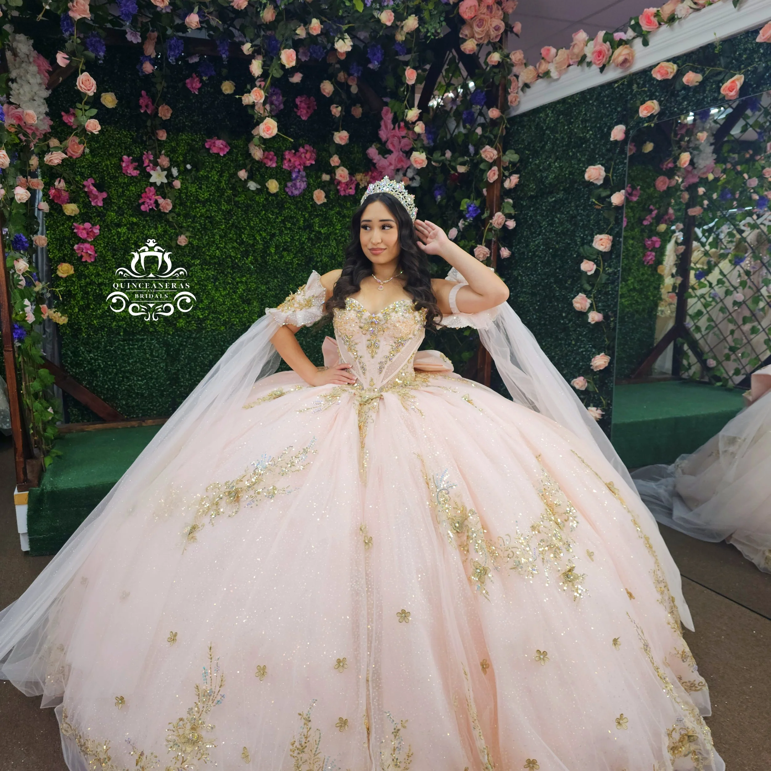 Young woman in a pink XV dress with gold embroidery, wearing a tiara, standing in front of a floral backdrop with pink and purple flowers, accessorized with jewelry and posing with one hand on her hip.