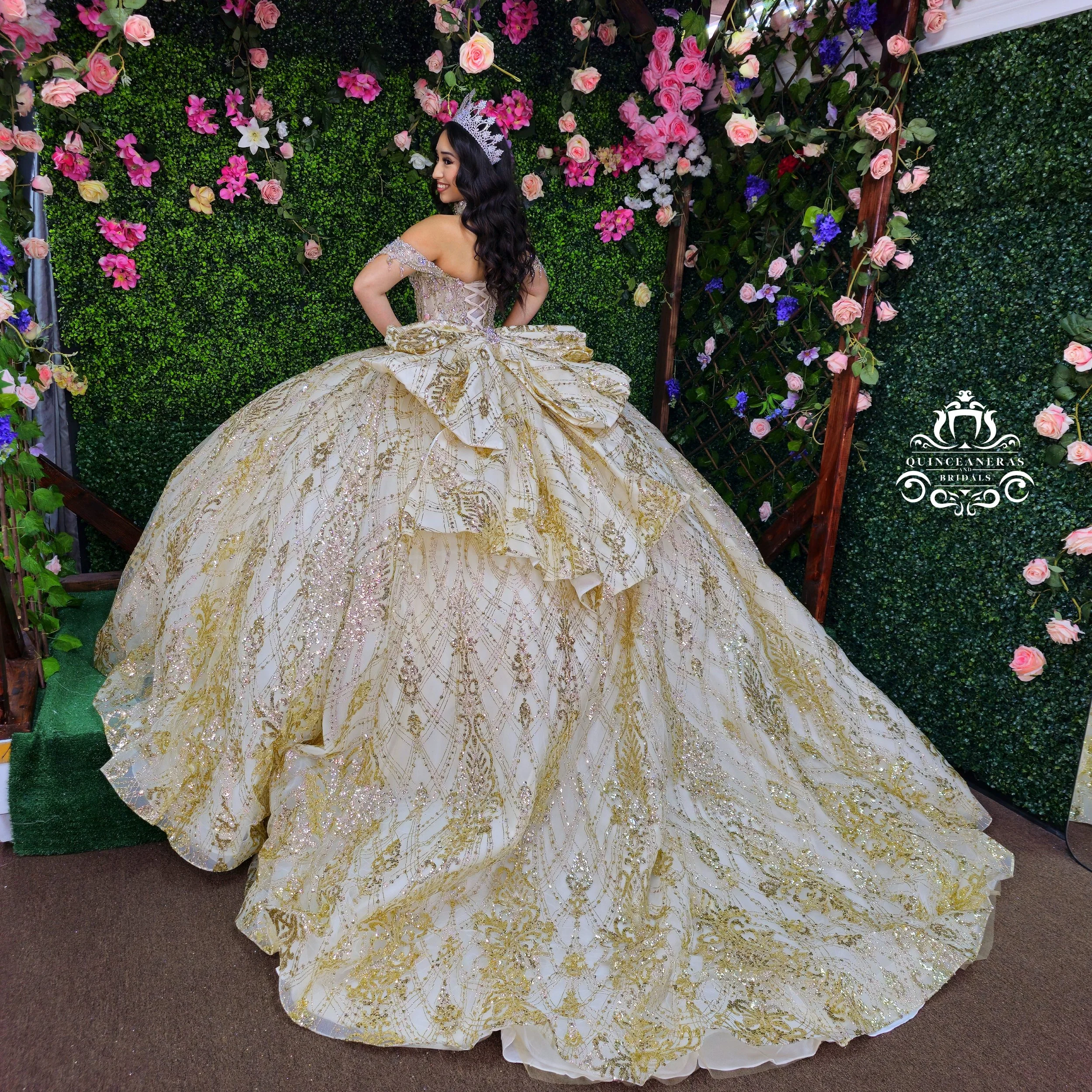 A woman in a gold and cream quinceanera dress with intricate embroidery and glitter, wearing a tiara, poses in front of a green hedge decorated with pink and purple flowers.