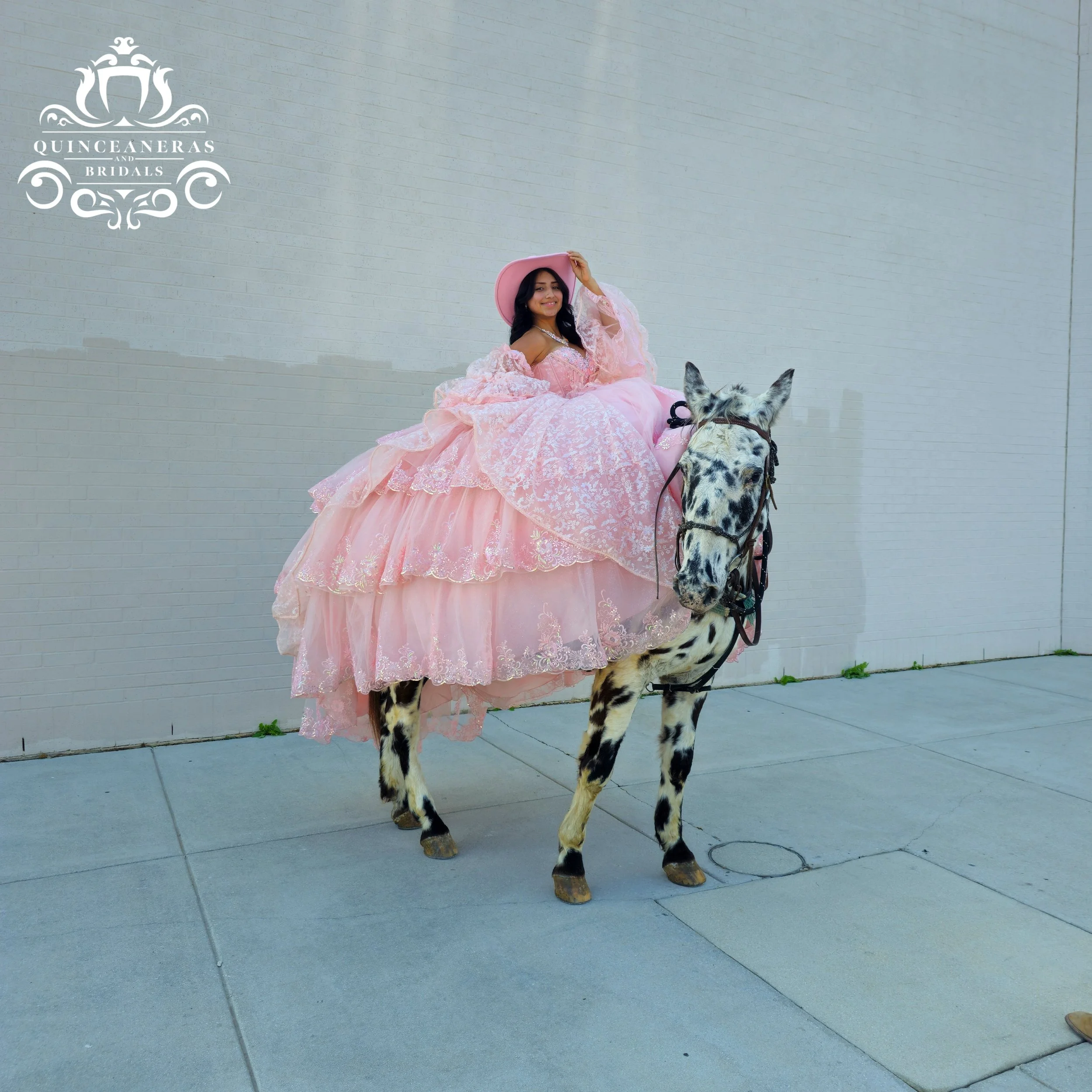 A woman in a pink, layered, lace western looking quince dress and a cowboy pink hat sits atop a spotted horse against a plain light-colored brick wall. The woman is smiling and holding her hat with one hand.