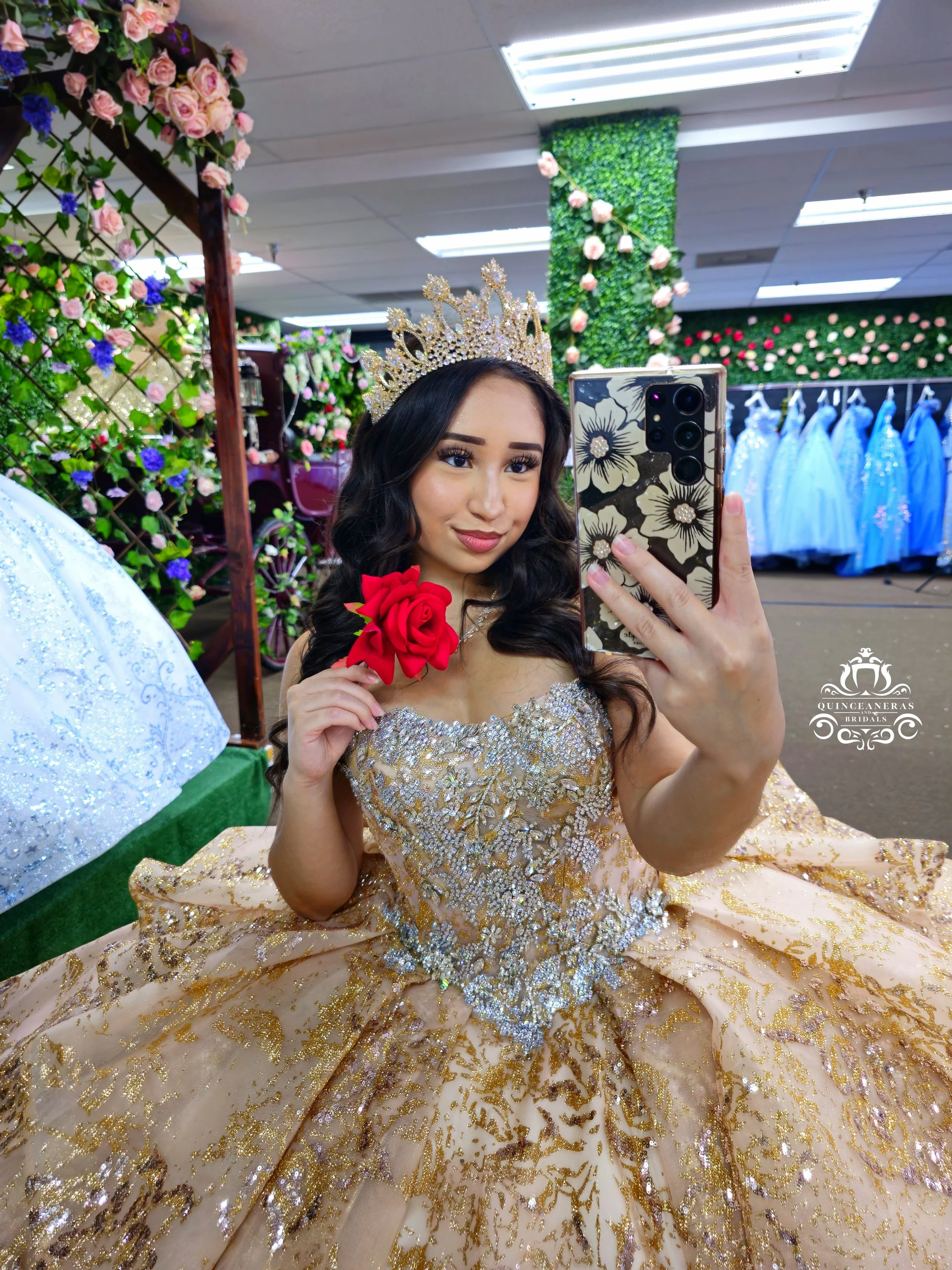 Young woman taking a selfie in a boutique with a princess XV gown, wearing a gold crown, holding a red rose, surrounded by floral decor and blue dresses in the background.