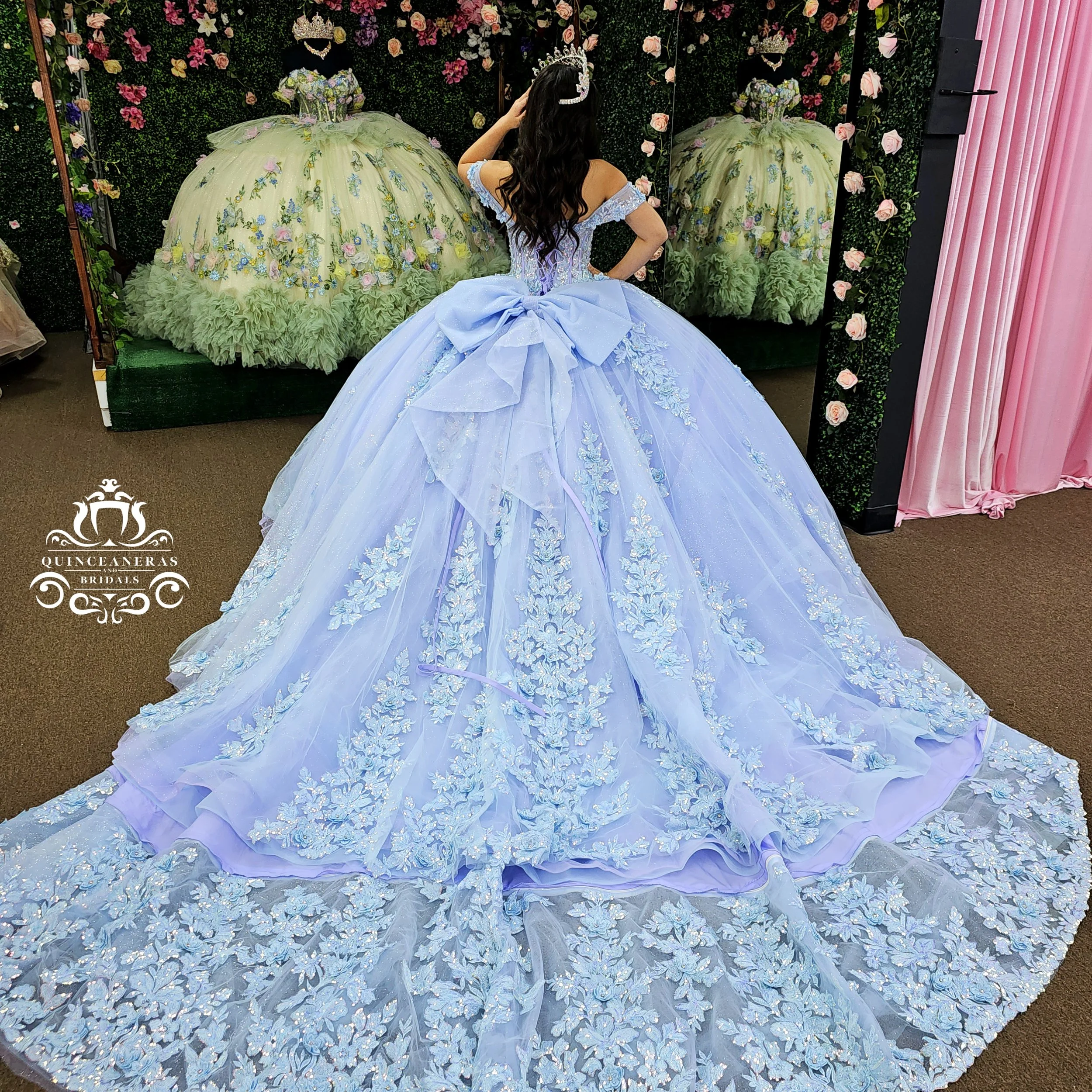 A woman wearing a light blue ball gown with floral embroidery and a large bow on the back, trying on the dress in a fitting room with pink curtains and floral decorations.