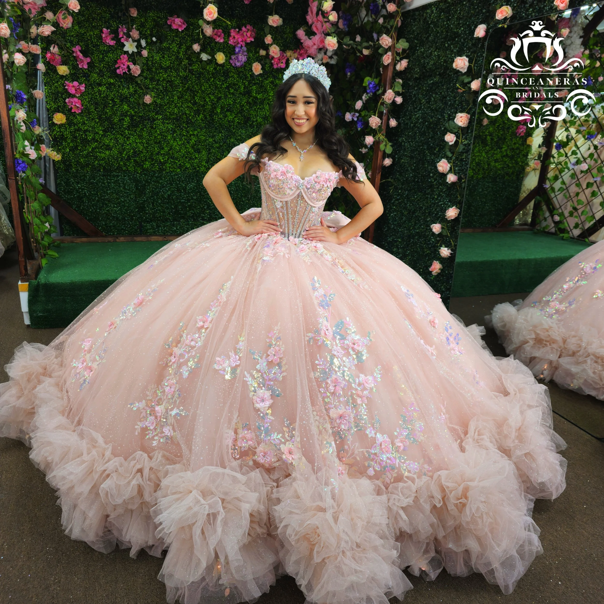 A young woman wearing a pink, floral-adorned ball gown with a full, ruffled skirt and off-the-shoulder neckline, standing in front of a floral green wall with a small stage and a sign that reads "Quinceañeras and Bridals." She is smiling and wearing 