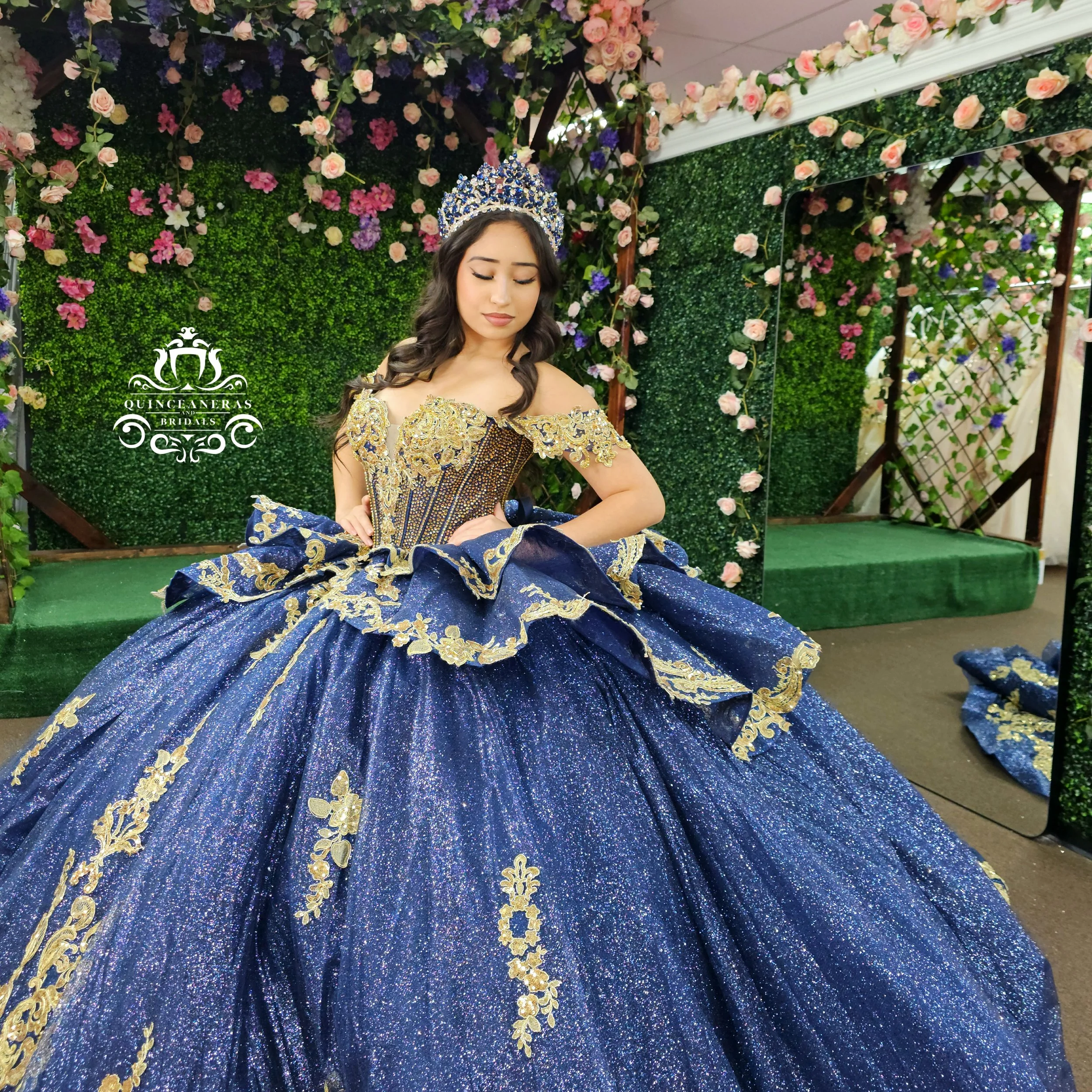 Young woman in a blue and gold quinceañera dress with a sparkling full skirt, standing in front of a floral backdrop, wearing a tiara, with her eyes closed.