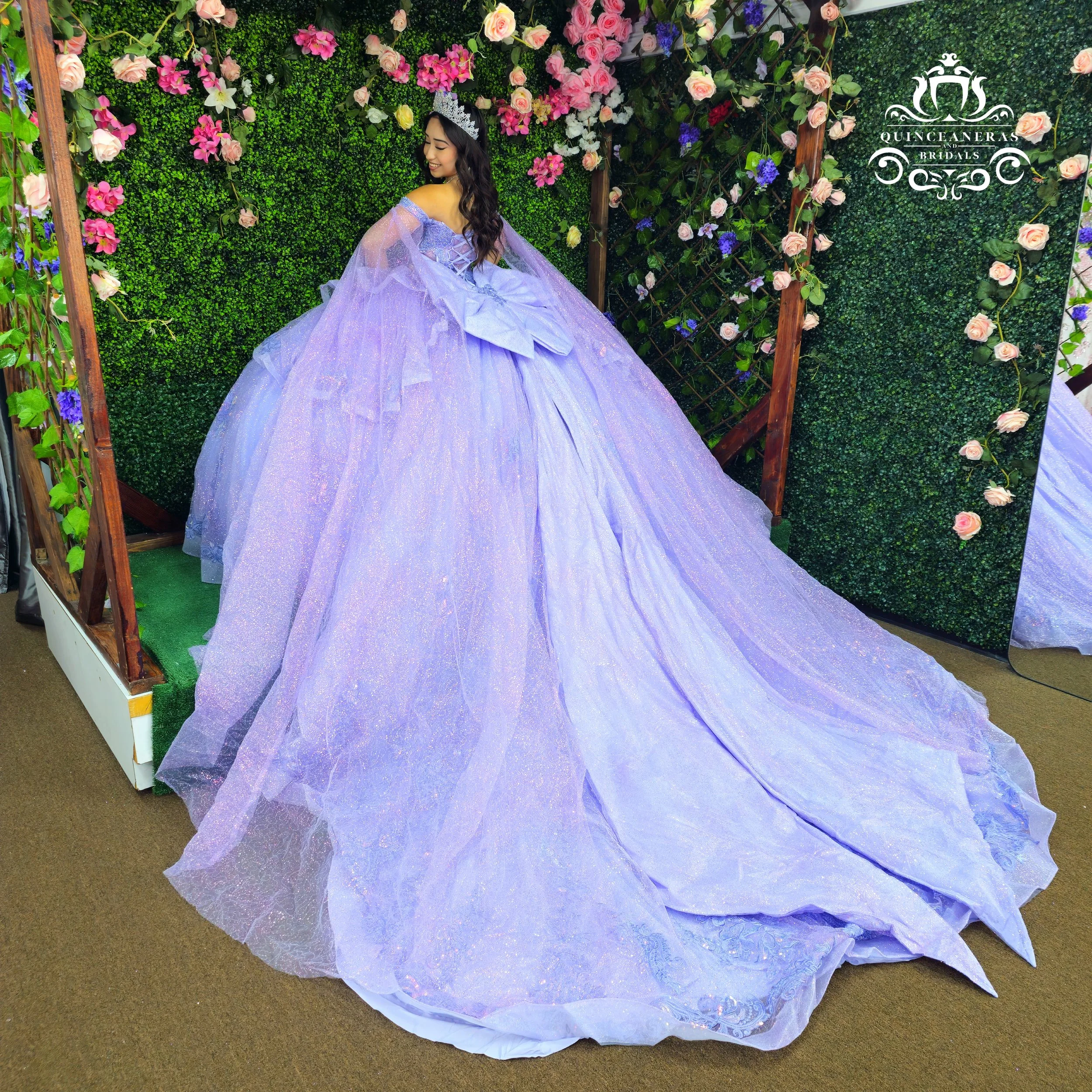 A young woman wearing a purple princess quinceanera dress and a tiara, standing in front of a decorative green wall with pink and purple flowers and smiling.