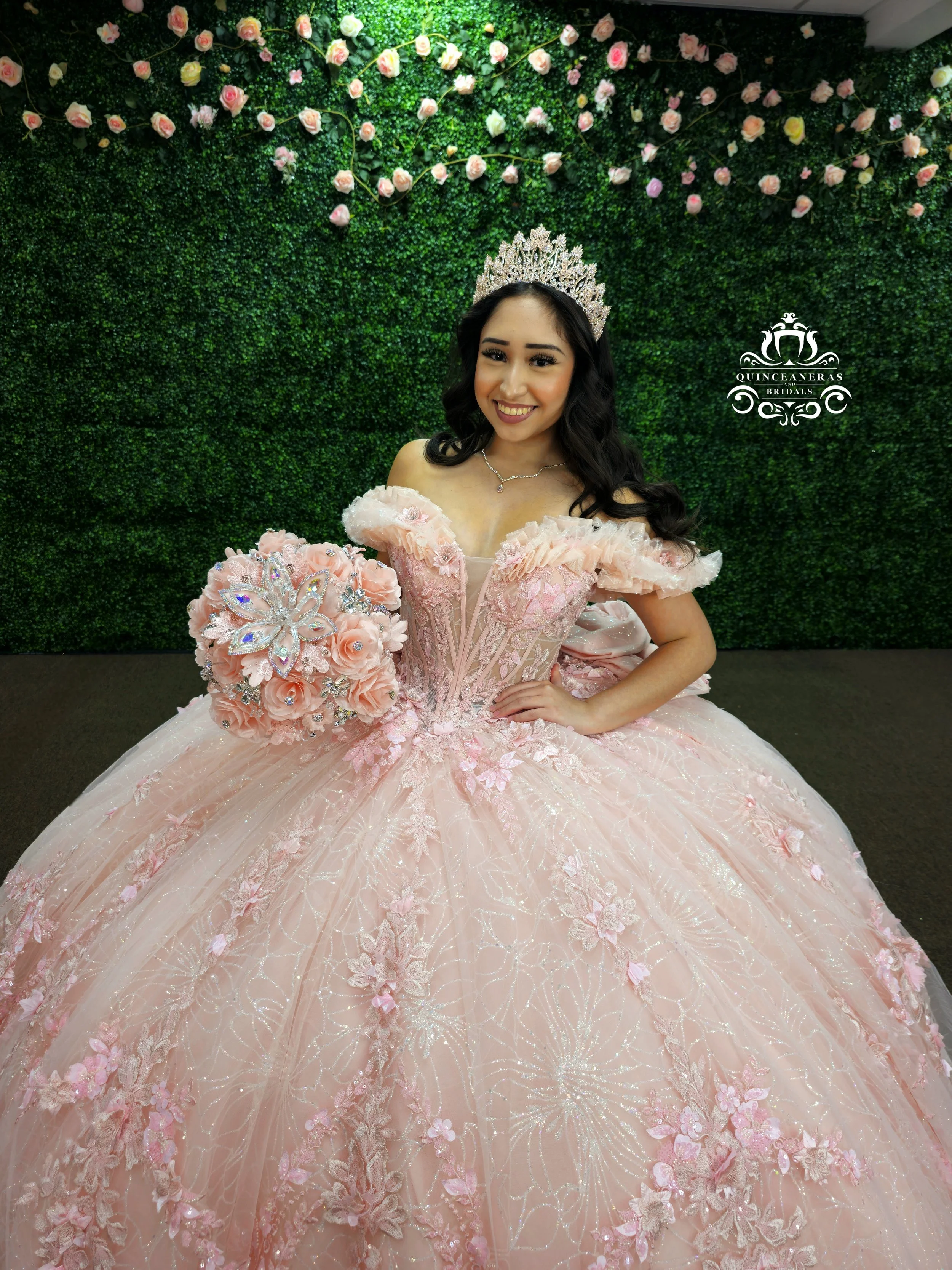 Young woman in a pink, floral, princess-style XV gown with off-shoulder sleeves, holding a bouquet of pink roses with a butterfly design, standing in front of a green floral wall with pink roses, wearing a silver tiara and necklace.