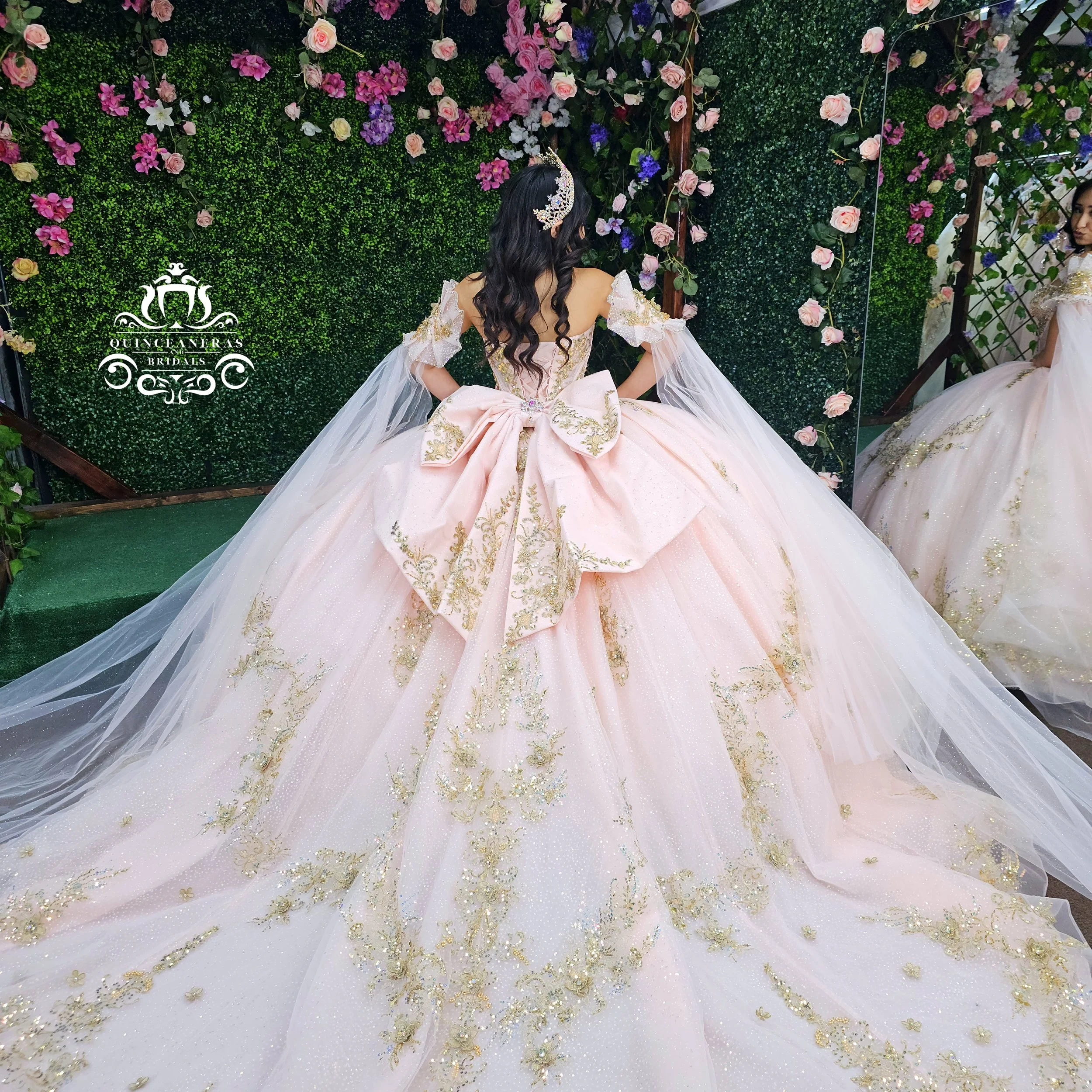 A woman in a pink quince dress with gold embroidery, an elaborate bow at the back, and off-the-shoulder tulle sleeves, standing before a green hedge wall decorated with pink, purple, and white flowers.