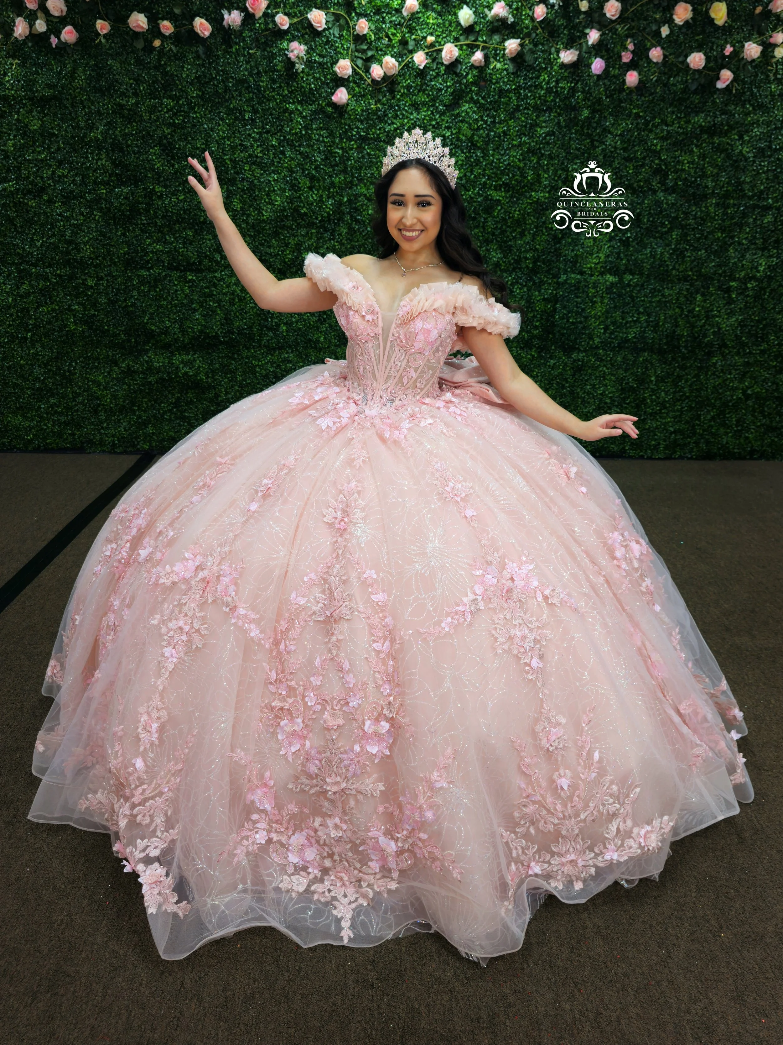 A woman in a pink, floral-embellished ball gown with an off-shoulder design and a tiara, standing in front of a green leafy backdrop with pink roses, smiling and posing with her arms raised.