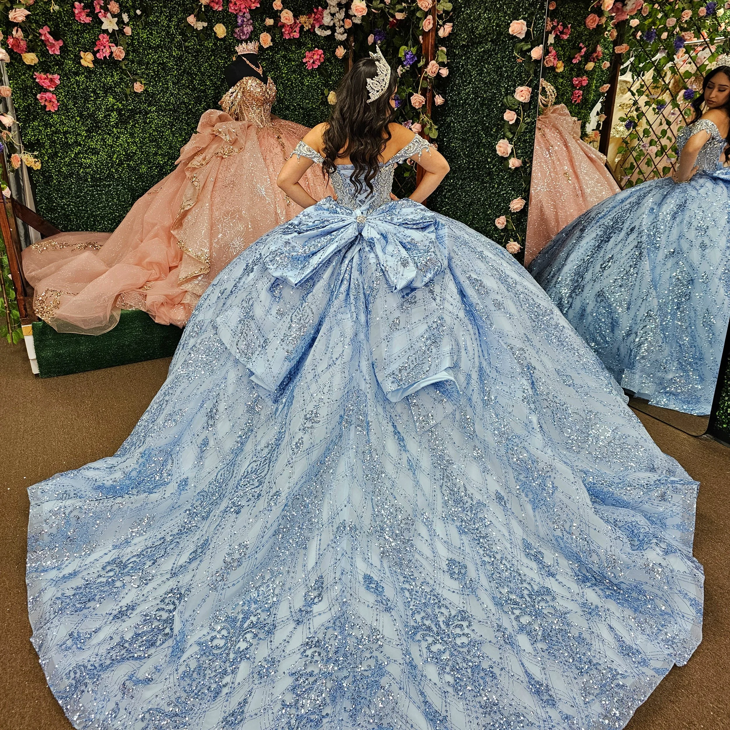 A woman trying on a large blue ball gown with a big bow on the back in a dress shop. The dress is decorated with silver glitter. There are other dresses and a floral wall in the background.