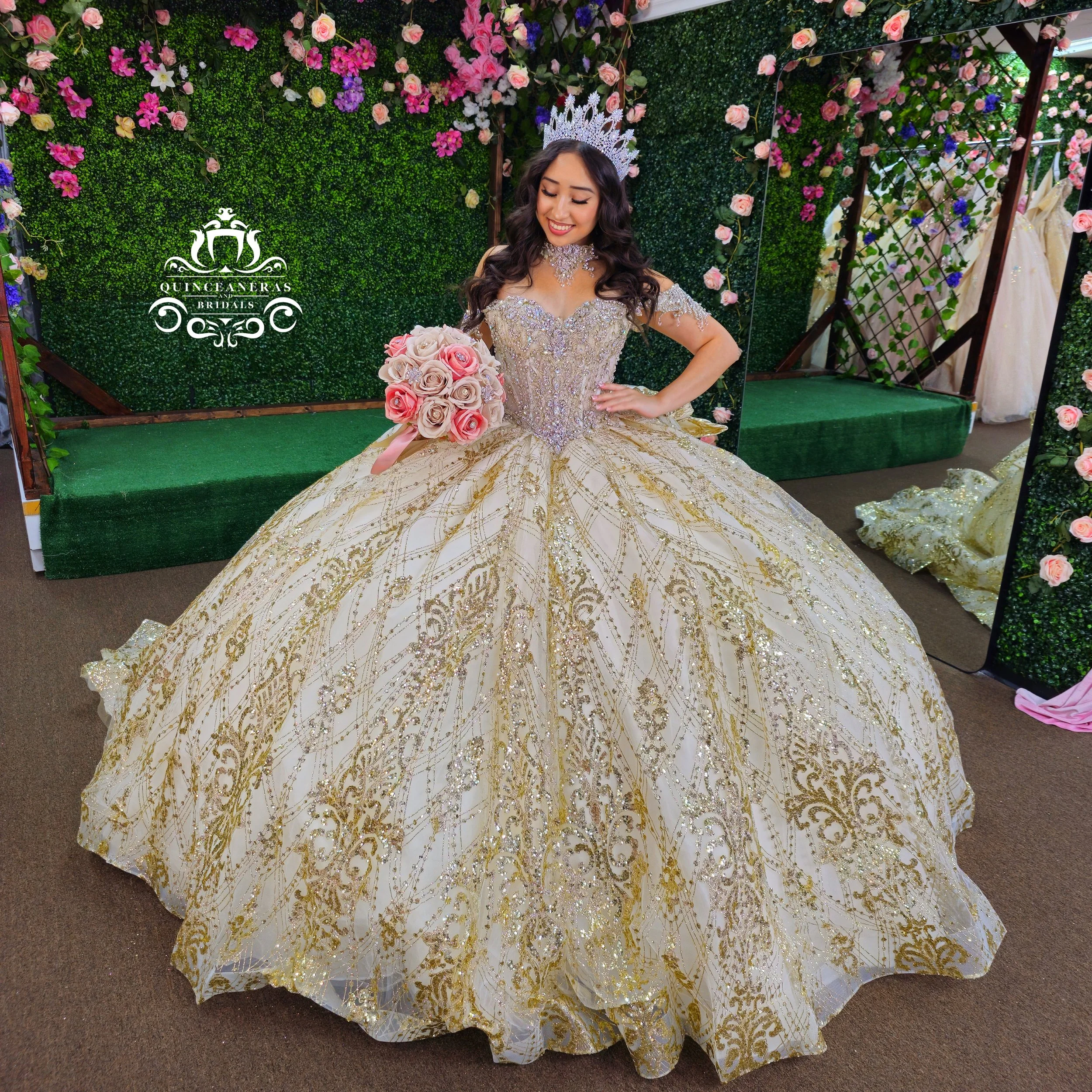 Quinceanera gown with gold and silver ornate embroidery, wearing a jeweled tiara, holding a bouquet of pink and light pink roses, smiling in a floral decorated backdrop with greenery and flowers. The setting appears to be Quinceaneras and Bridals.