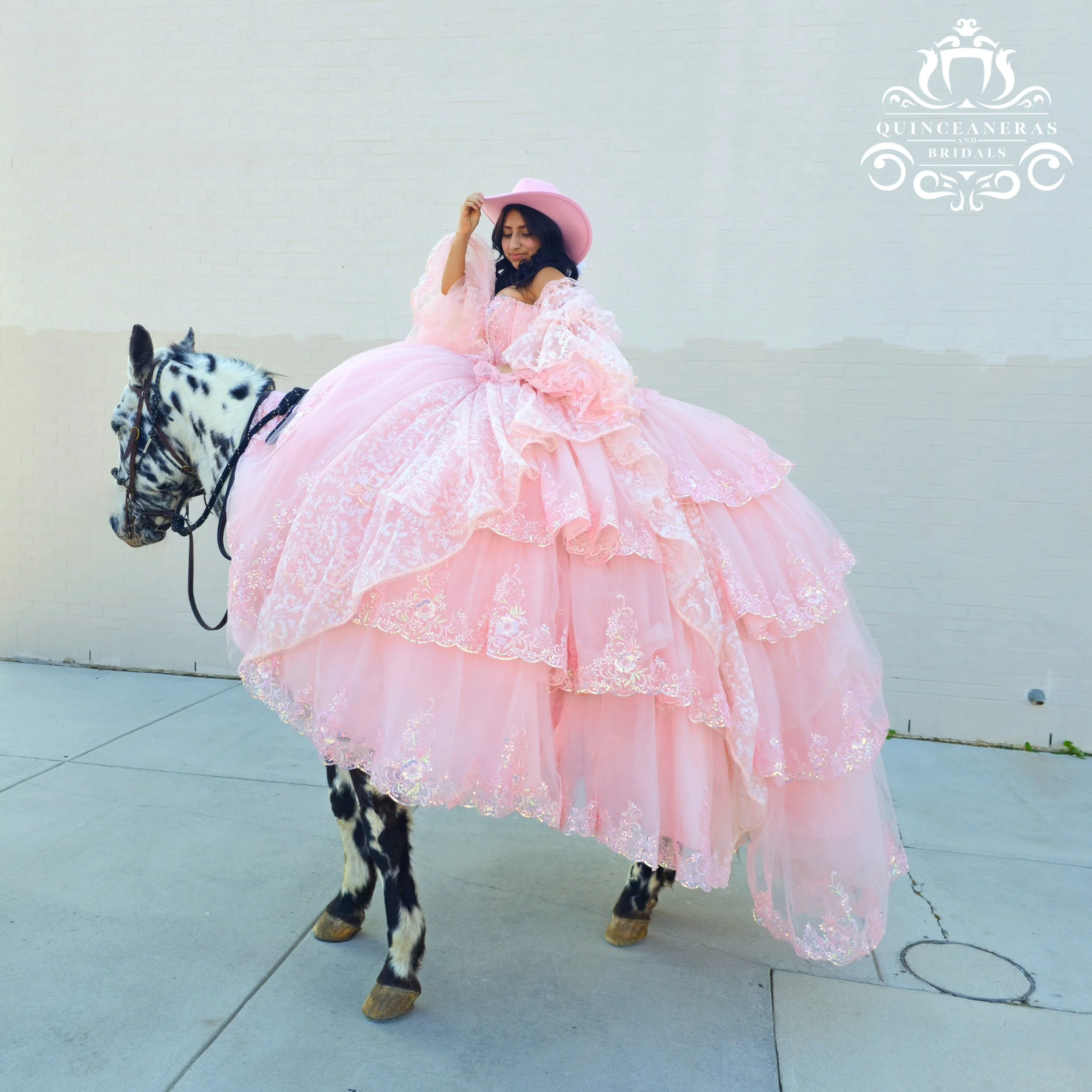 A woman in a voluminous pink XV dress and matching hat riding a black and white Spotted Saddle horse with a white face. a very western theme is present. 