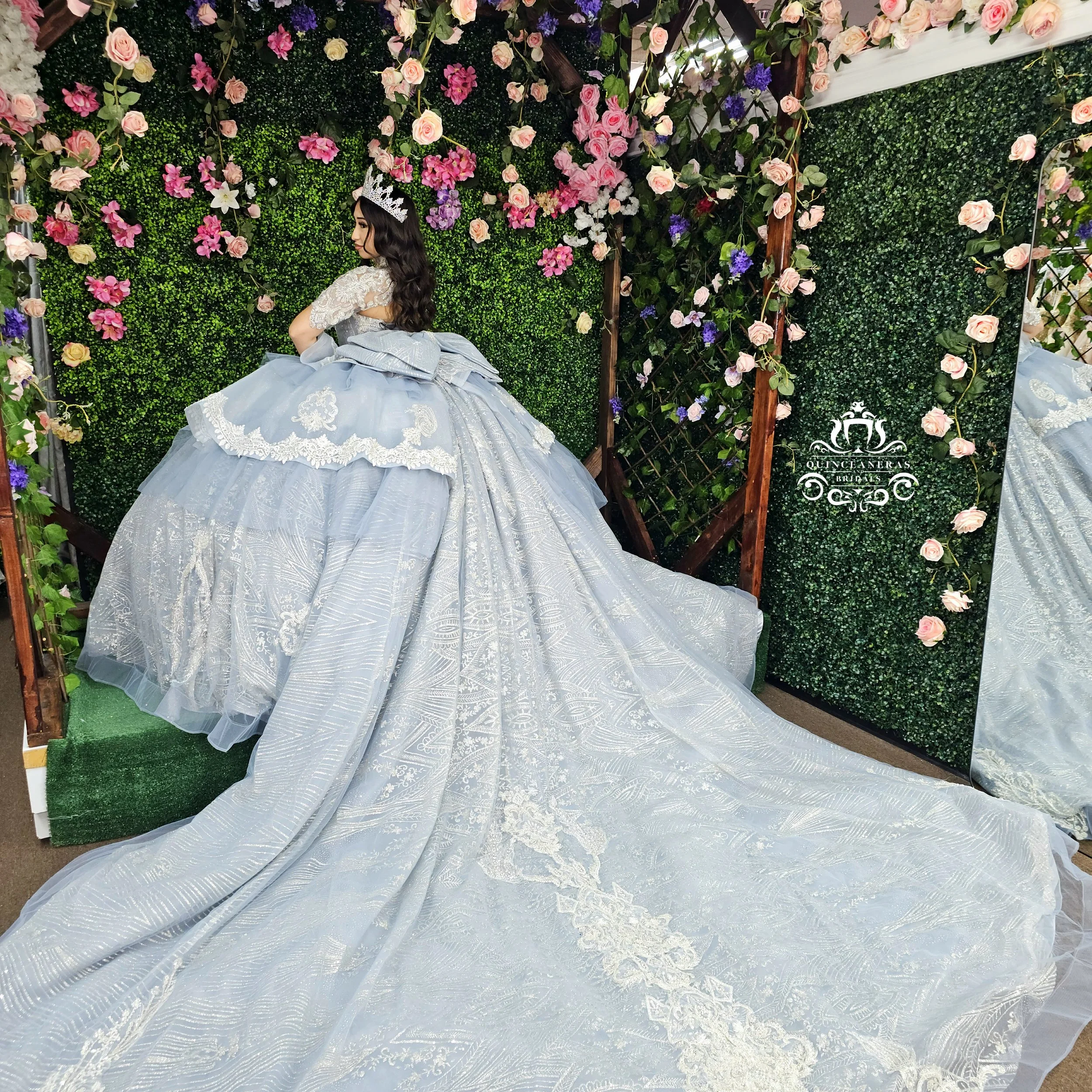 A woman wearing a light blue ball gown with lace details and a large train, standing in front of a green hedge wall decorated with pink, purple, and white flowers, and a mirror on the right side.