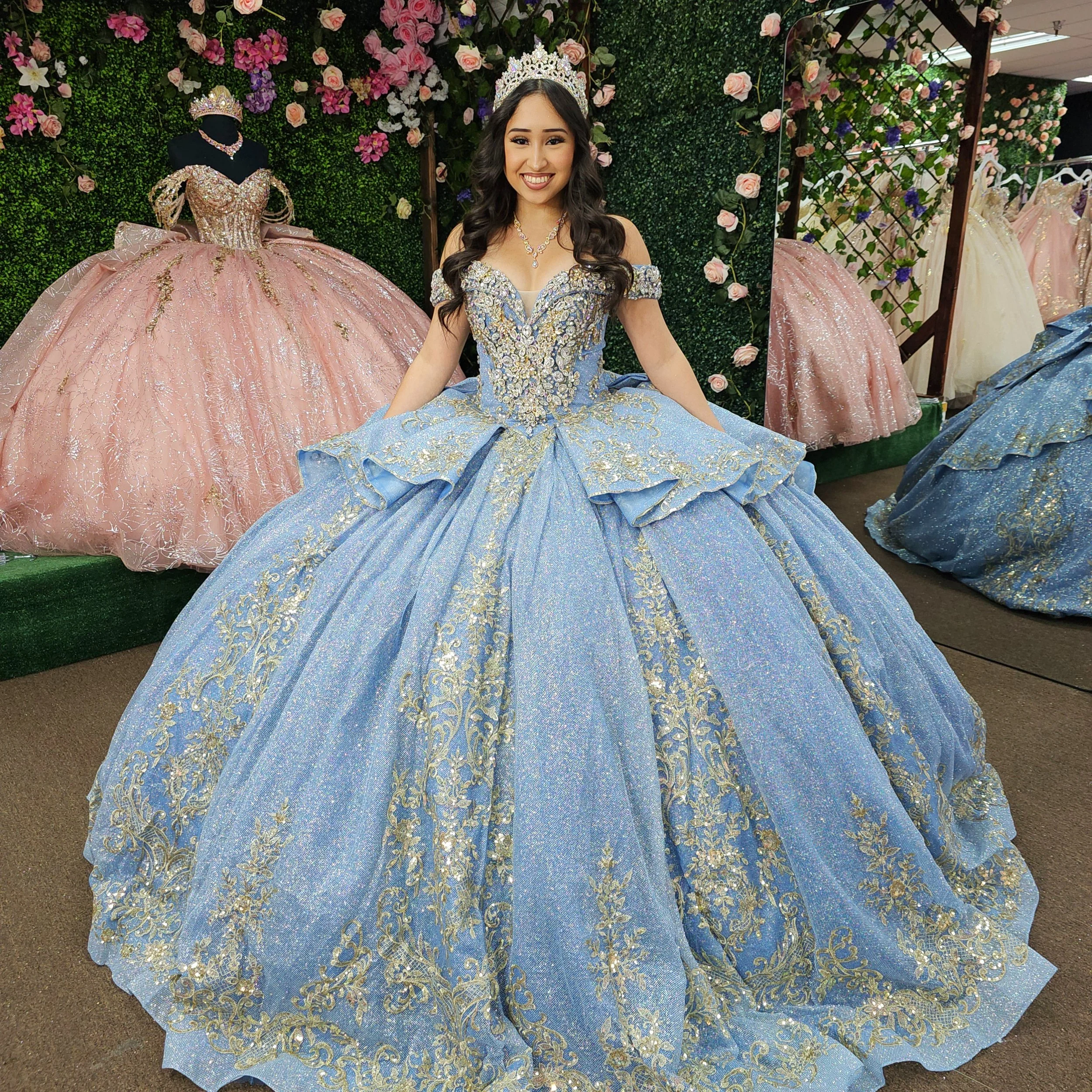 Young woman in a blue and gold ornate ball gown, wearing a tiara, standing in front of pink and purple floral display with other elaborate dresses in the background.