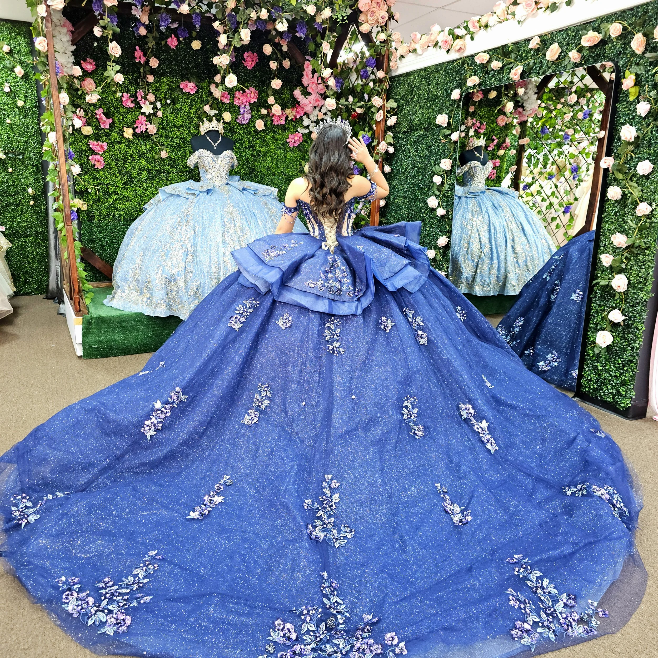 A woman trying on a blue xv ball gown with floral embroidery, standing in front of a large mirror, surrounded by other embellished gowns and floral decorations.
