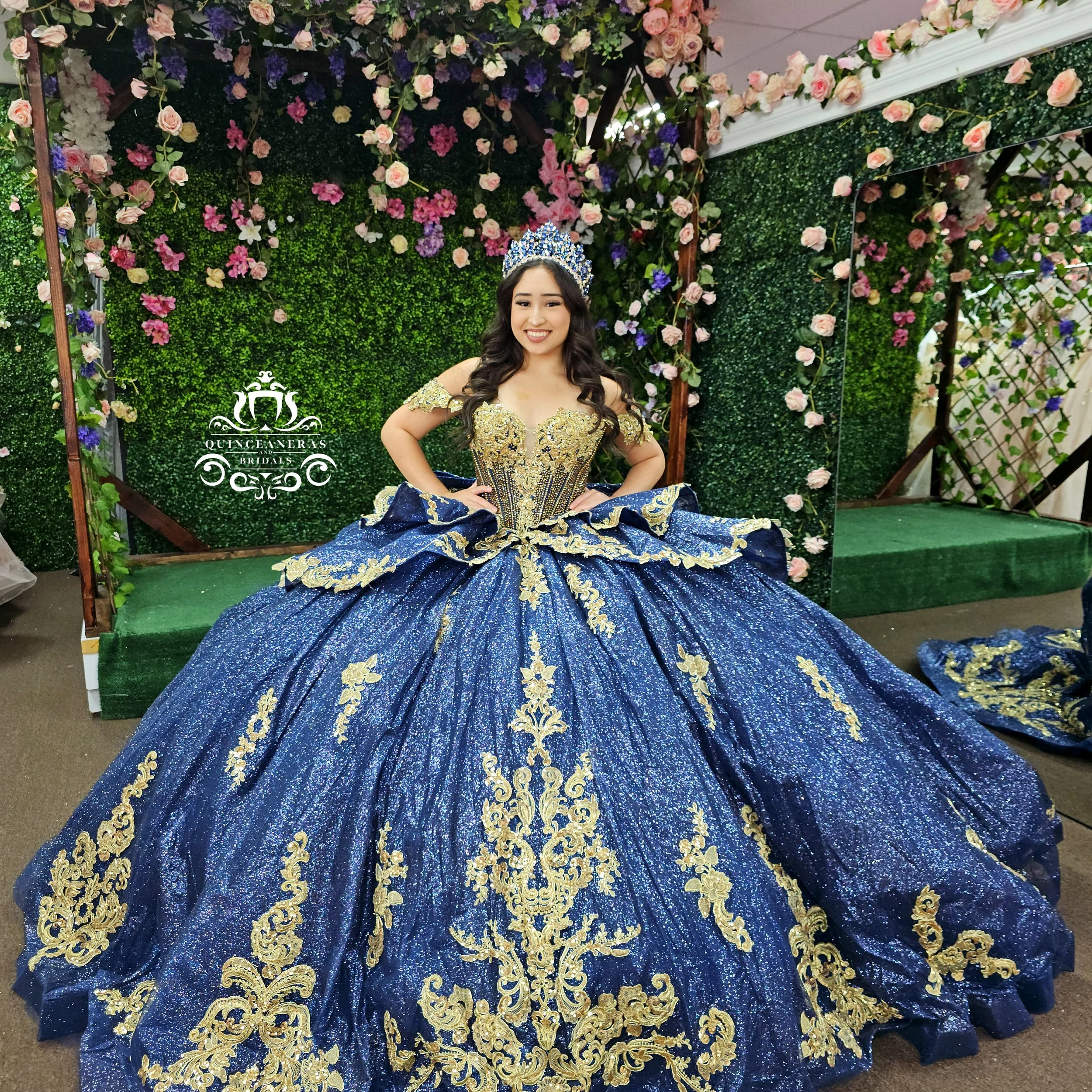 A young woman wearing a royal blue ball gown with gold embroidery, a tiara, and smiling at a floral backdrop.
