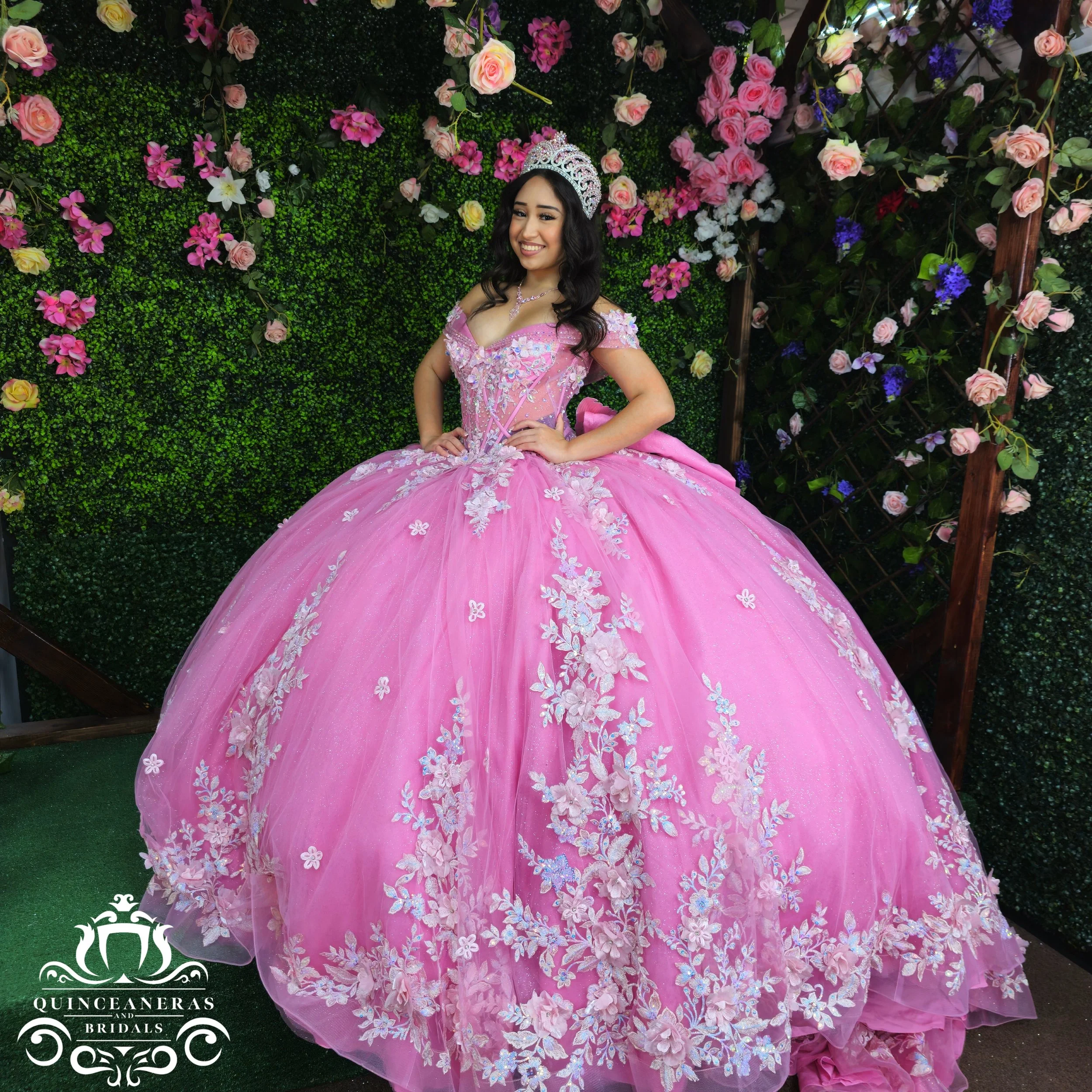Young woman in a pink quince dress with floral embroidery, wearing a tiara, smiling, standing in front of a green wall decorated with pink, purple, and white flowers.