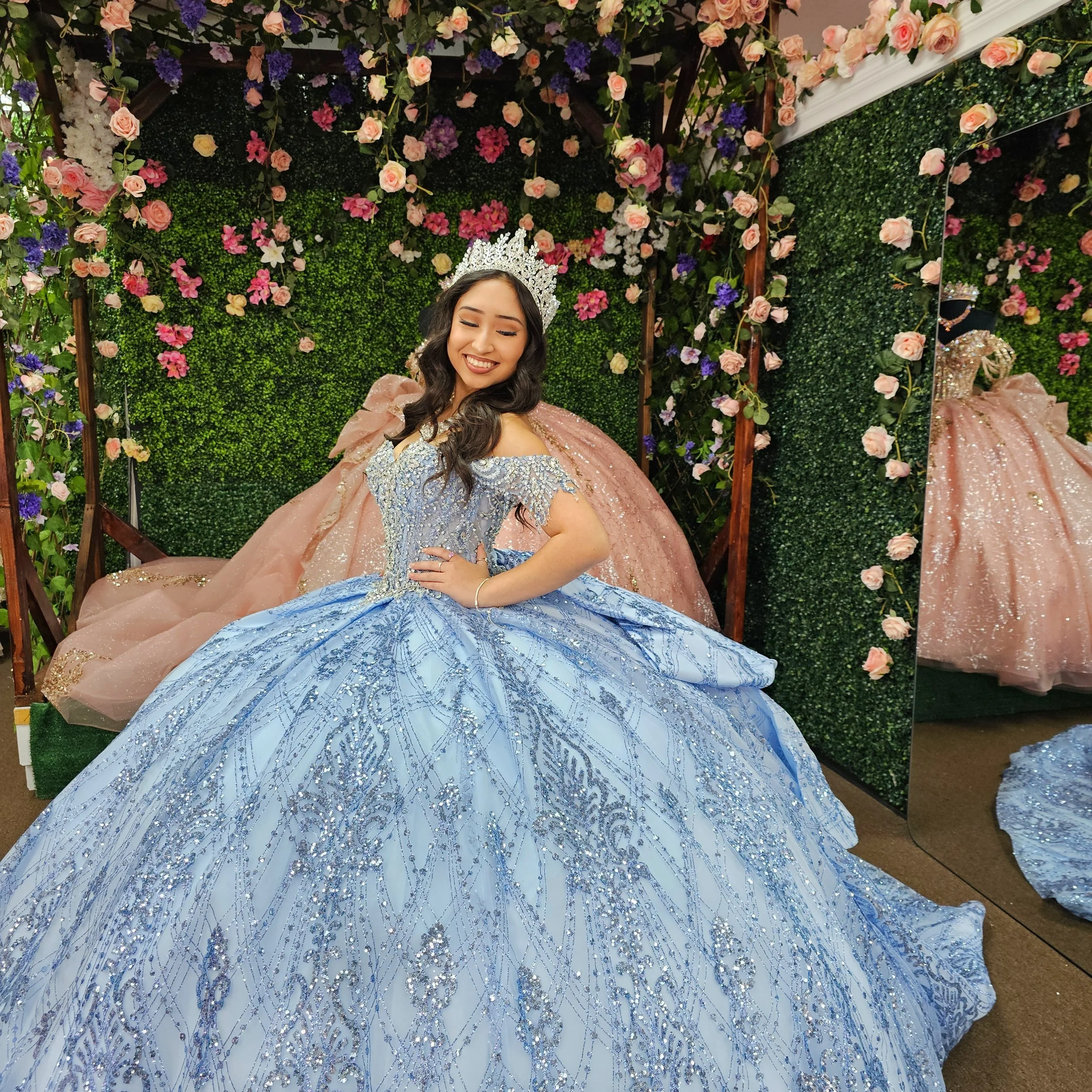 A young woman in a light blue quinceanera ball gown with intricate silver and blue embroidery, wearing a sparkling tiara, is smiling and posing with her hand on her hip in a floral decorated boutique.