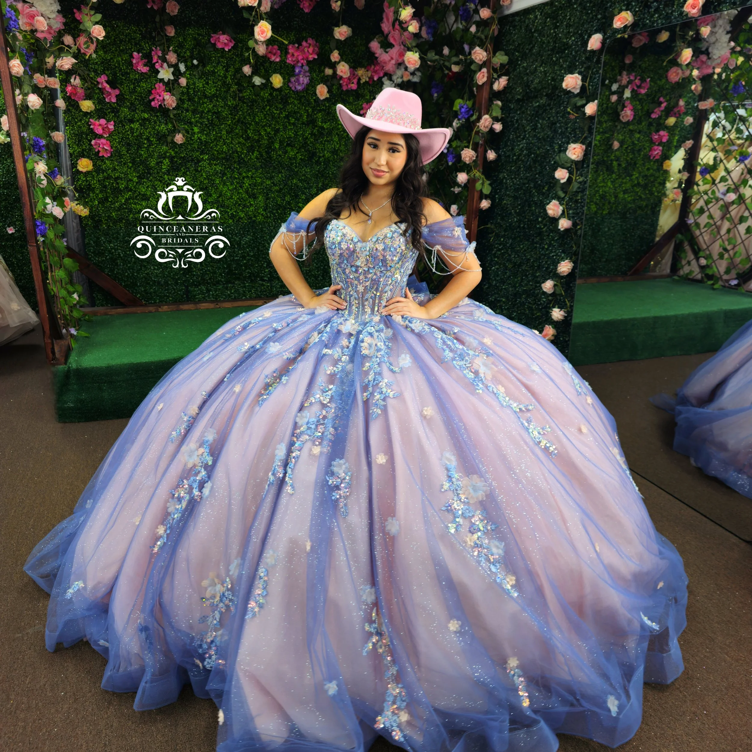 Young woman in a pastel purple and blue ball gown with floral embroidery, wearing a pink cowboy hat, standing in front of a floral and greenery backdrop at a quinceañera event.