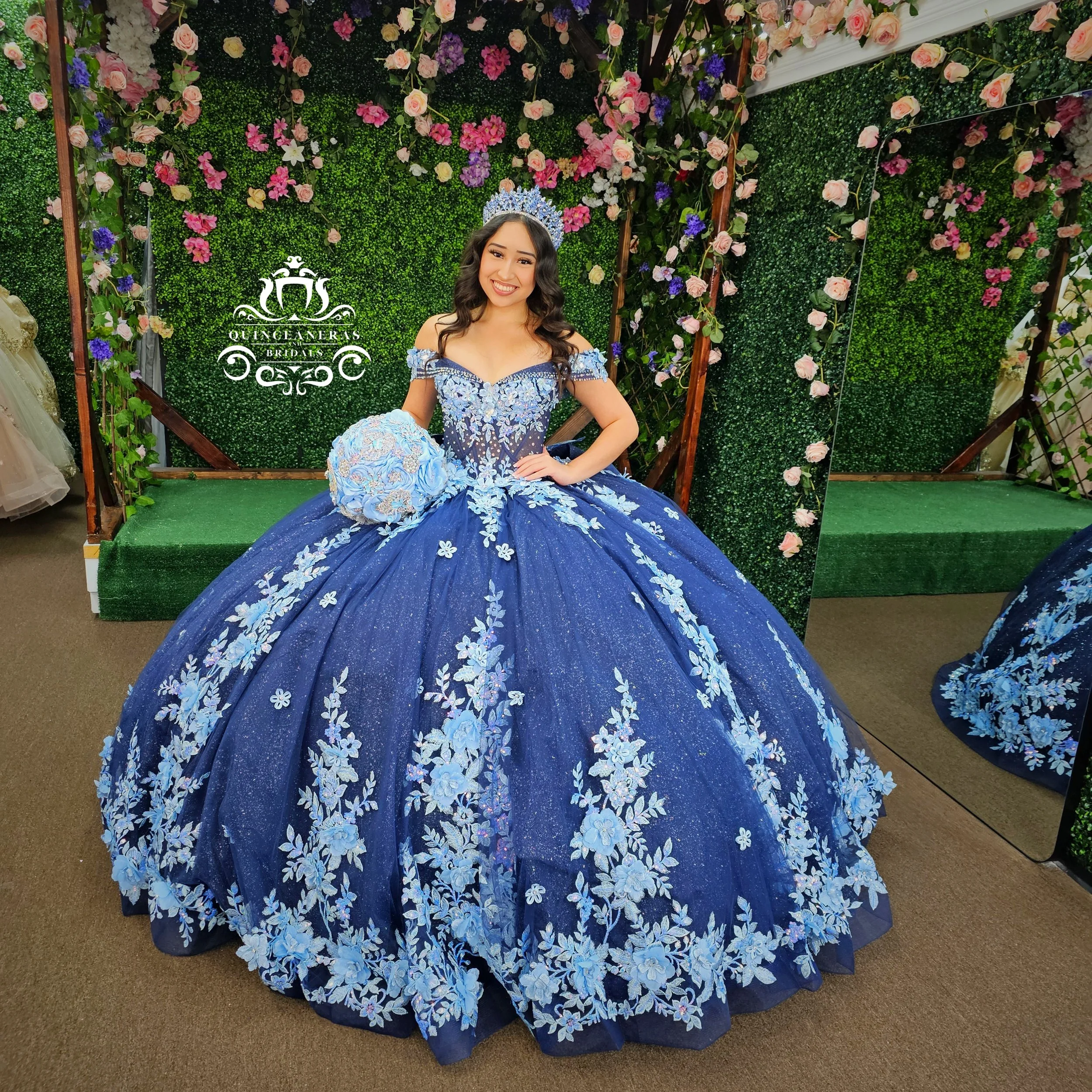 Young woman in a blue floral wedding gown with off-shoulder sleeves, standing in front of a green floral backdrop with pink and purple flowers, wearing a tiara and holding a matching bouquet.