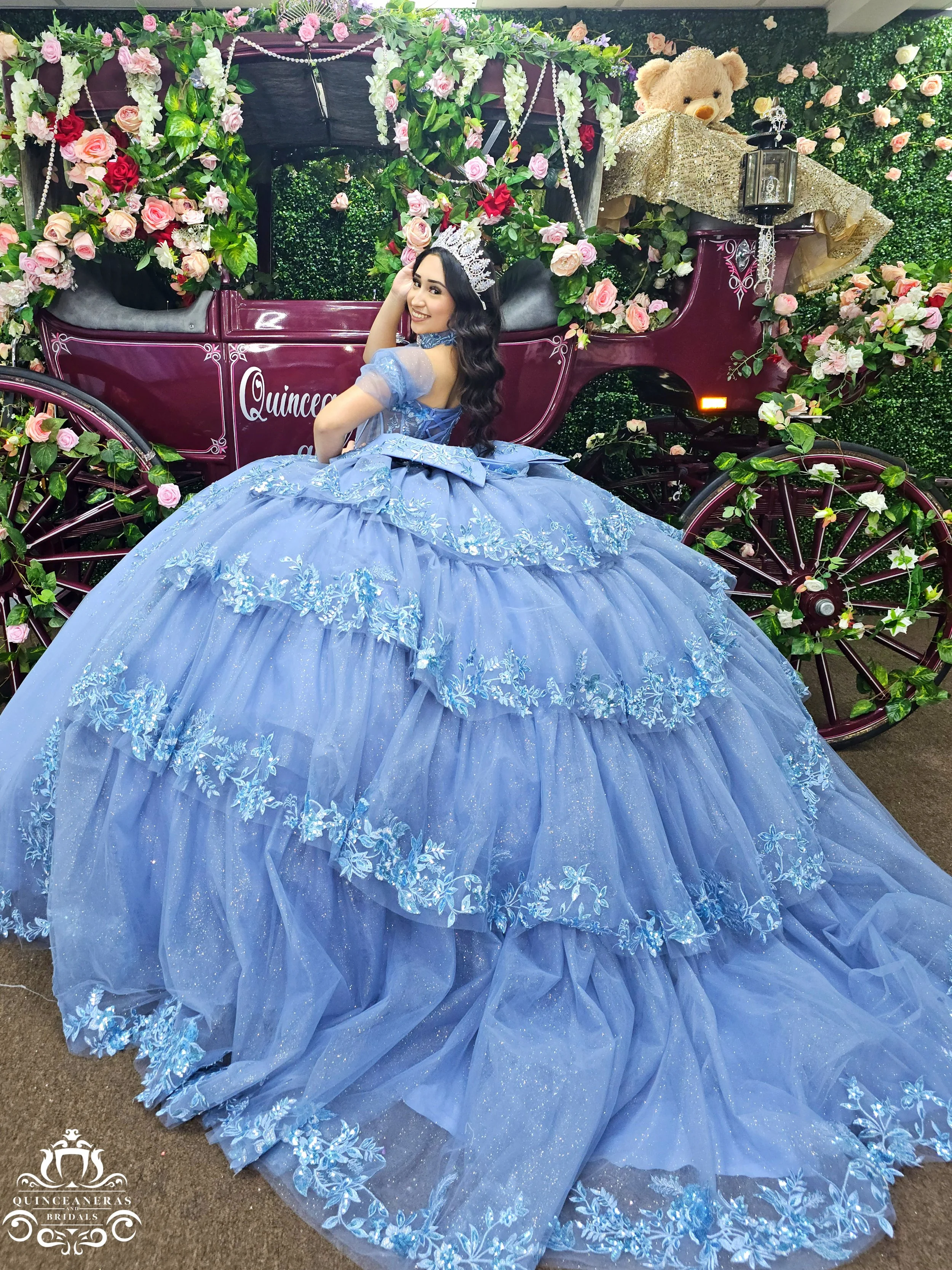 Youth girl in a blue princess dress with a silver tiara, smiling, sitting on a decorated carriage surrounded by pink and white roses, with a teddy bear on top of the carriage.