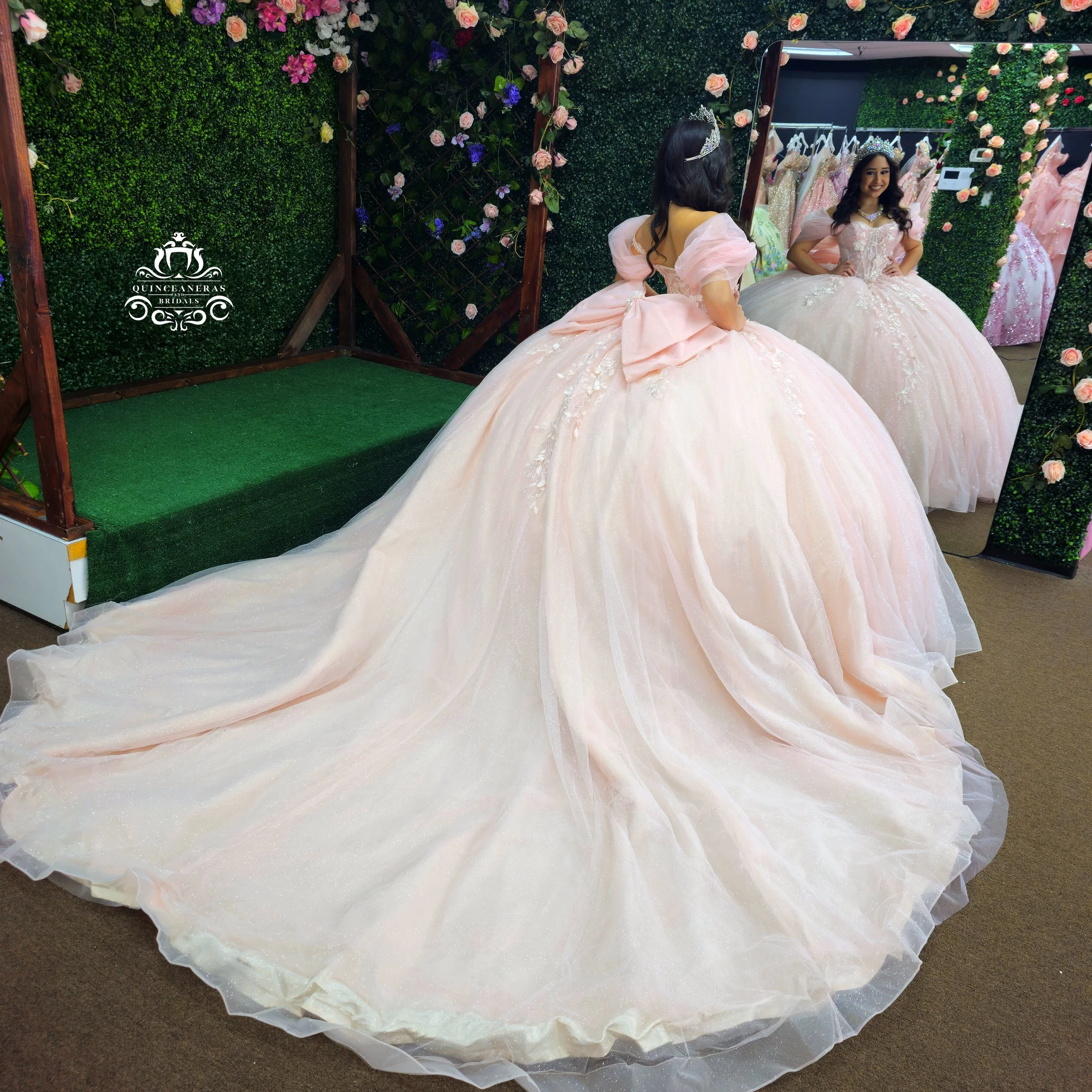 A woman trying on a pink, ball gown quince dress with a long train and puffy sleeves, looking at herself in a mirror.