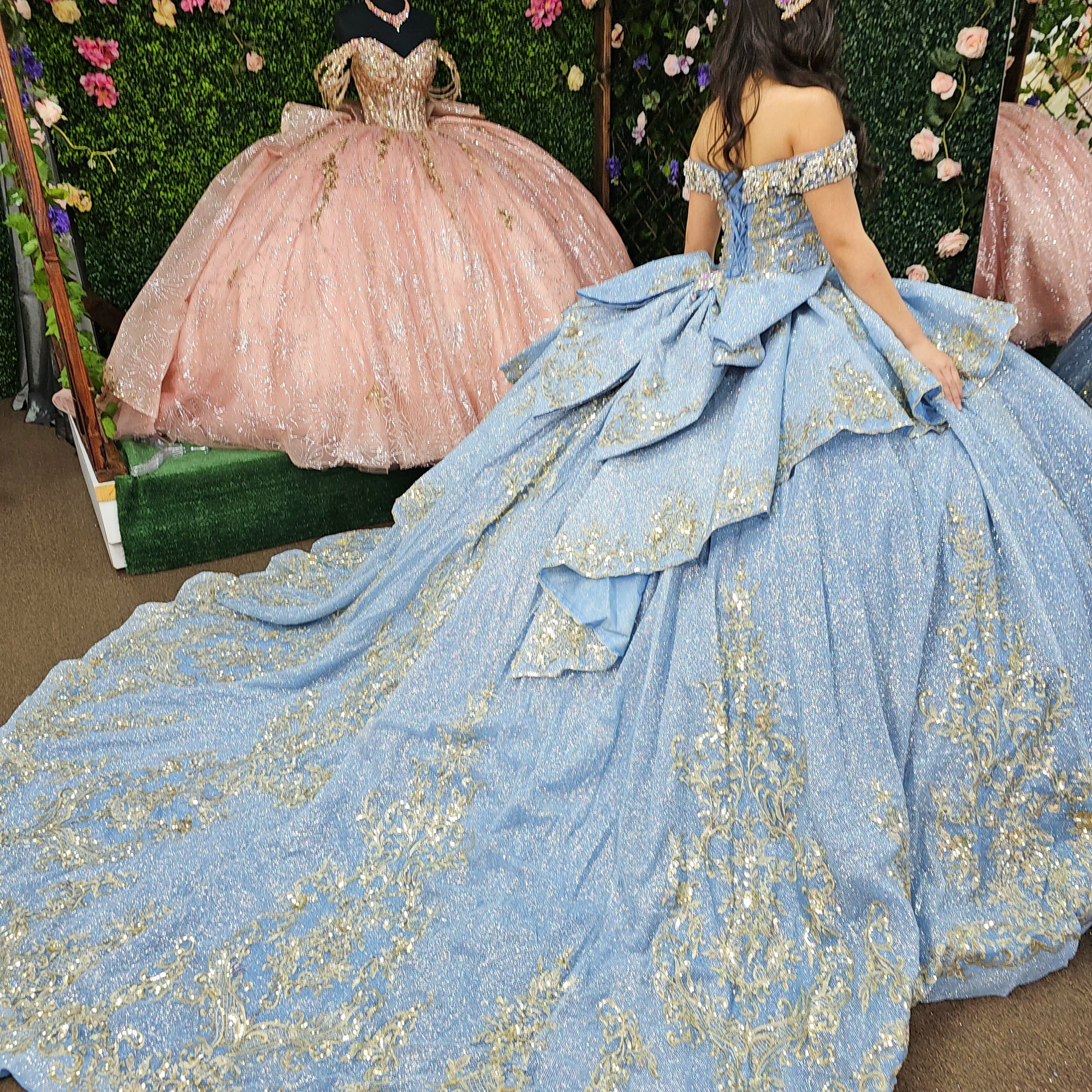 A woman trying on a sparkling blue gown with intricate gold details and a large back bow, in front of pink and pastel-colored ball gowns and a floral backdrop.