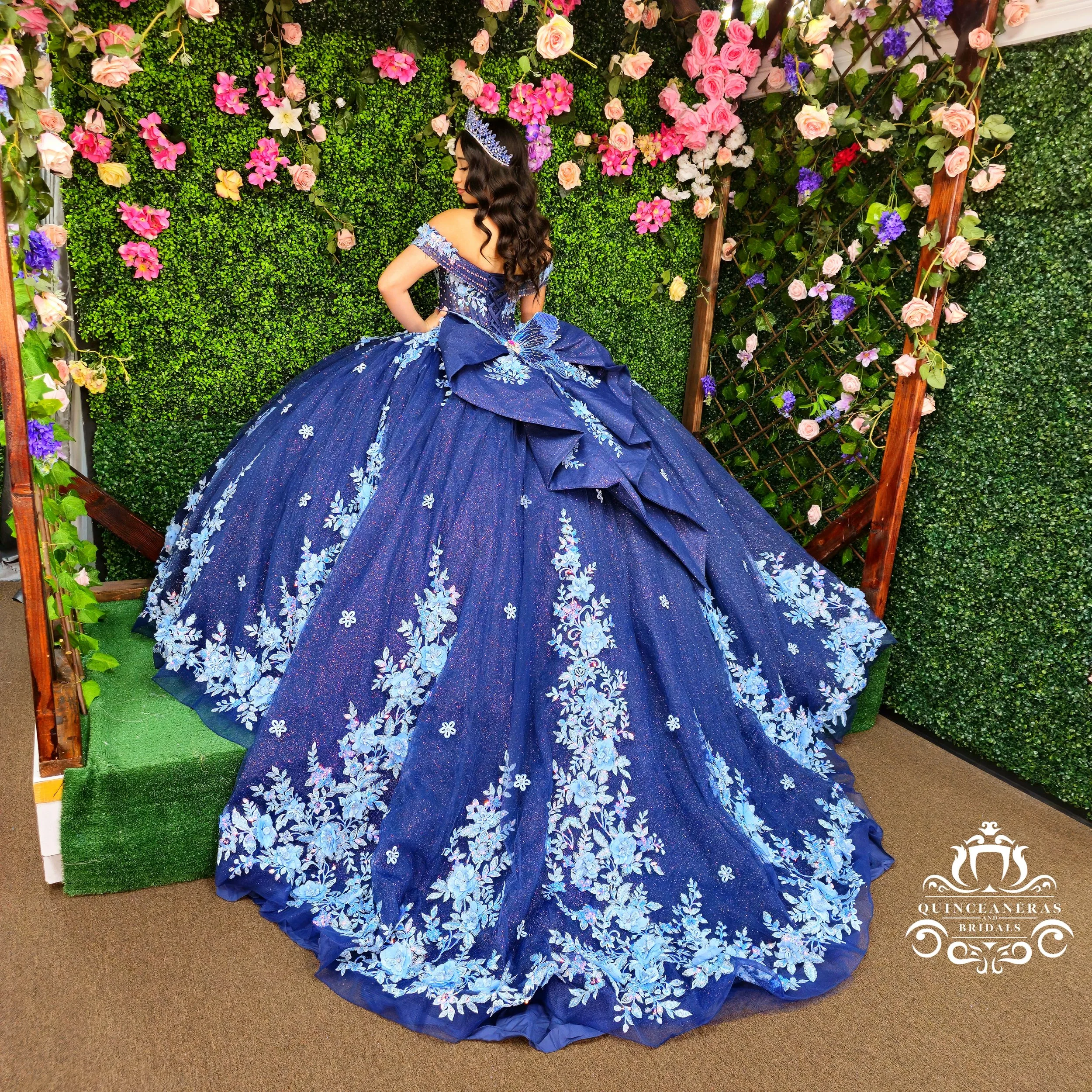 A woman in a blue, floral-patterned ball gown with off-the-shoulder sleeves and a large back bow, standing in front of a green leafy floral wall with pink and purple flowers. She has long, wavy dark hair and wears a tiara.