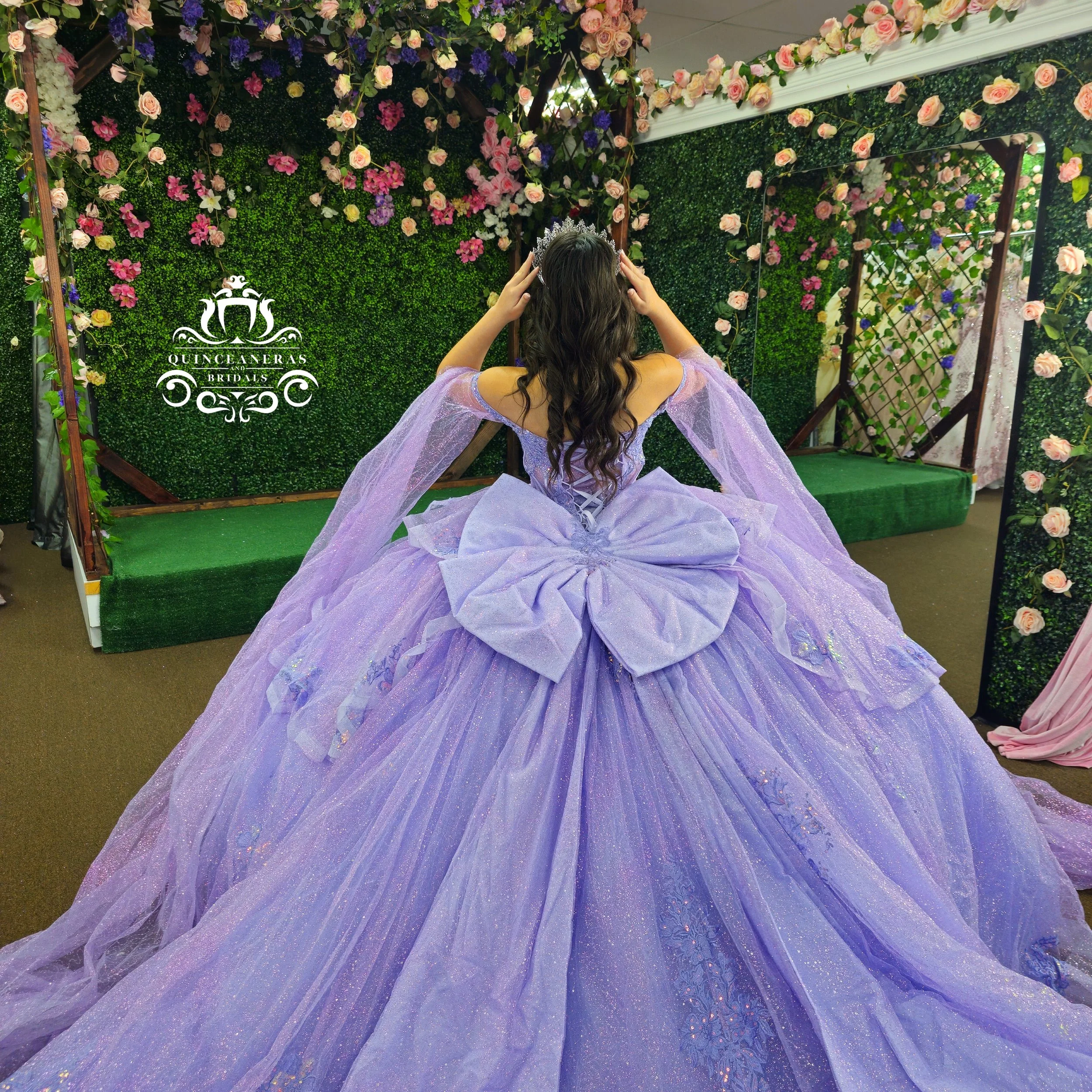 A woman in a lavender quince ball gown with a large bow on the back, posing in front of a floral backdrop with greenery, pink, purple, and yellow flowers, and a mirror.