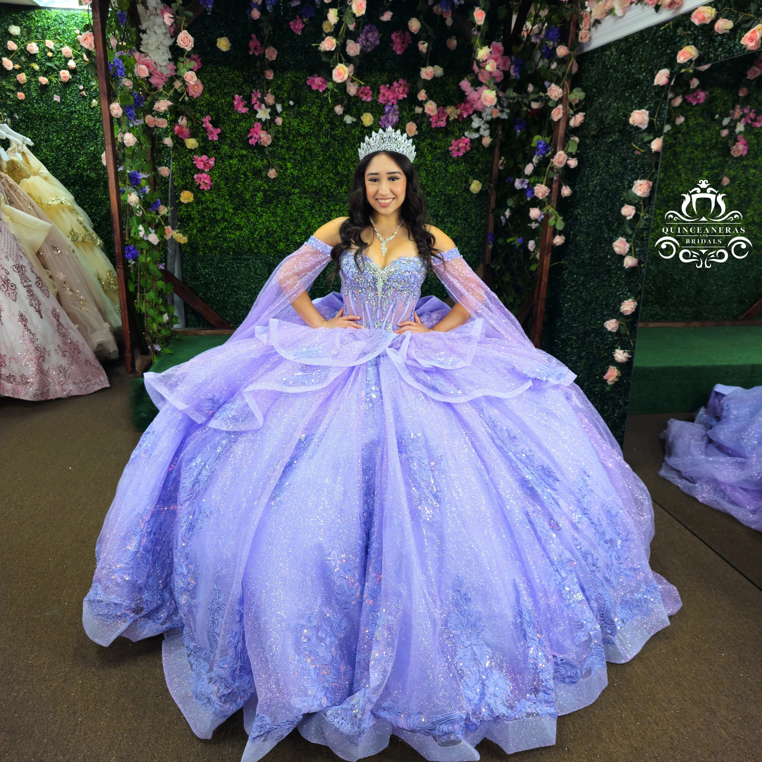 A woman in a lavender quinceañera gown with a tiara standing in front of a floral backdrop.