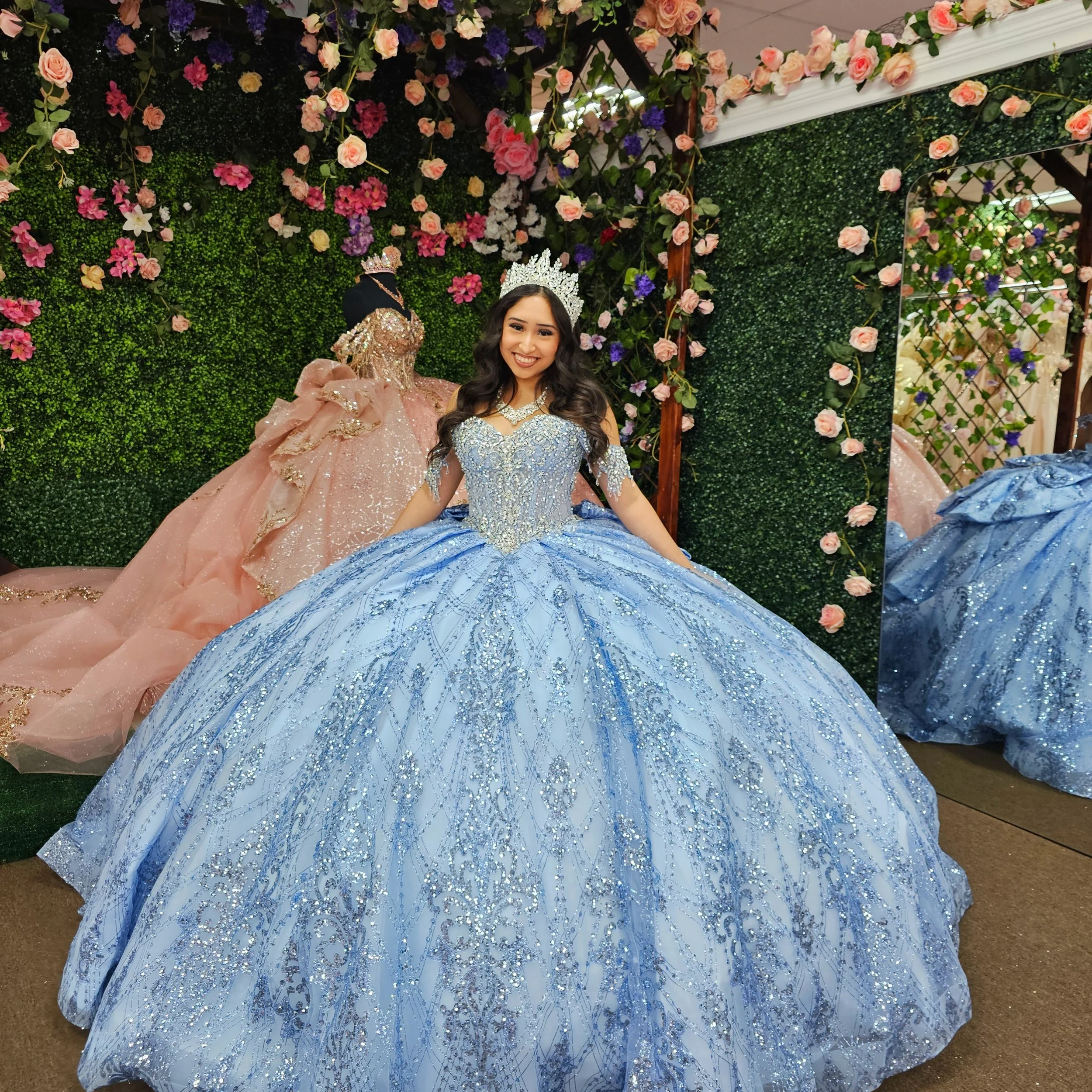 A young woman in a glittery blue quince ball gown with silver embellishments, wearing a tiara, smiling, in front of a floral backdrop.