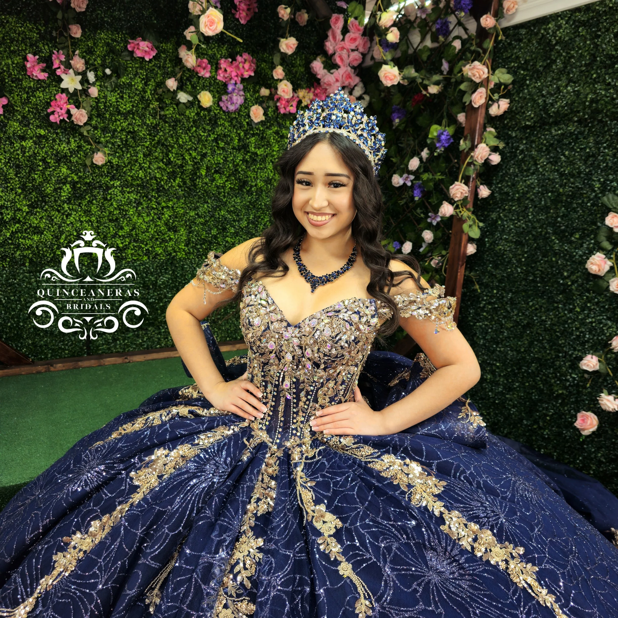 Young woman wearing a fancy dark blue and gold gown and a crown, sitting in front of a green leafy wall with pink, purple, and white flowers.