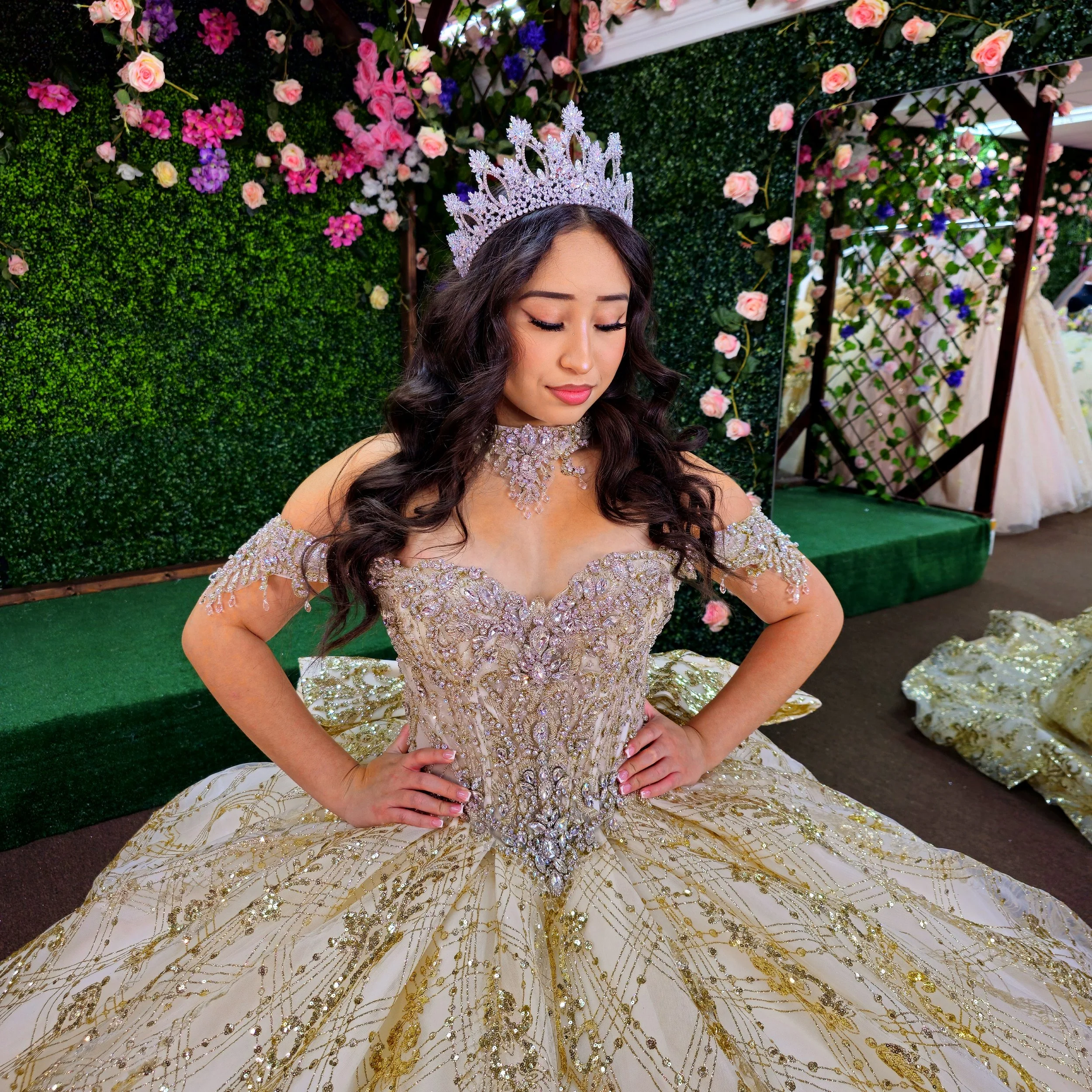Young woman in a gold, sparkling quince dress and a silver tiara, kneeling with hands on her hips, on a decorated green background with flowers at a celebration or event.