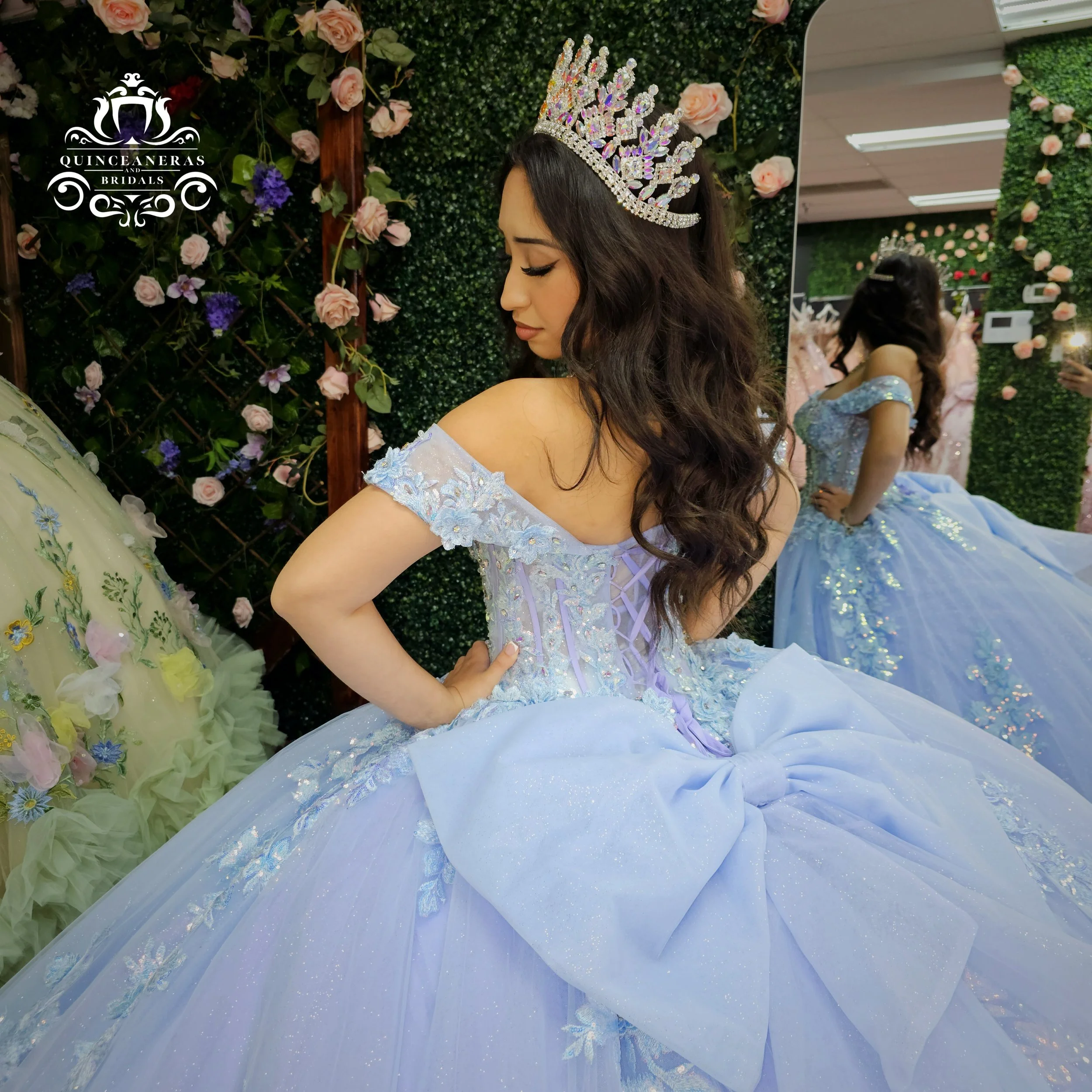 A young woman trying on a lavender ball gown wedding dress and a sparkling crown at a bridal shop. She is standing with her hand on her hip while looking down, and her reflection is visible in a mirror behind her, with a background of green foliage a