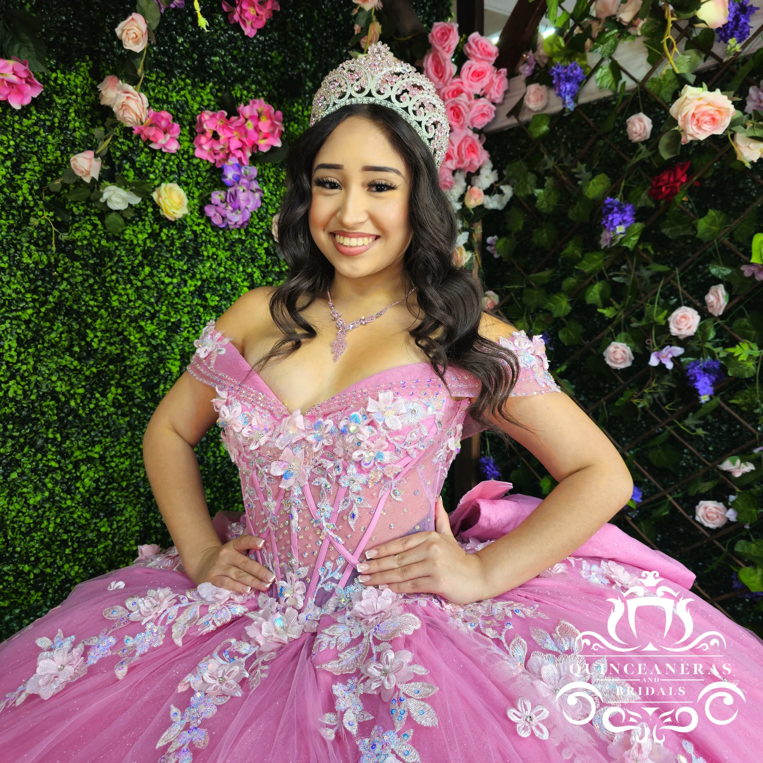 A young woman in a pink quinceañera gown with floral embroidery, lace, and bead embellishments, wearing a silver tiara and matching necklace, standing in front of a lush green and floral backdrop.