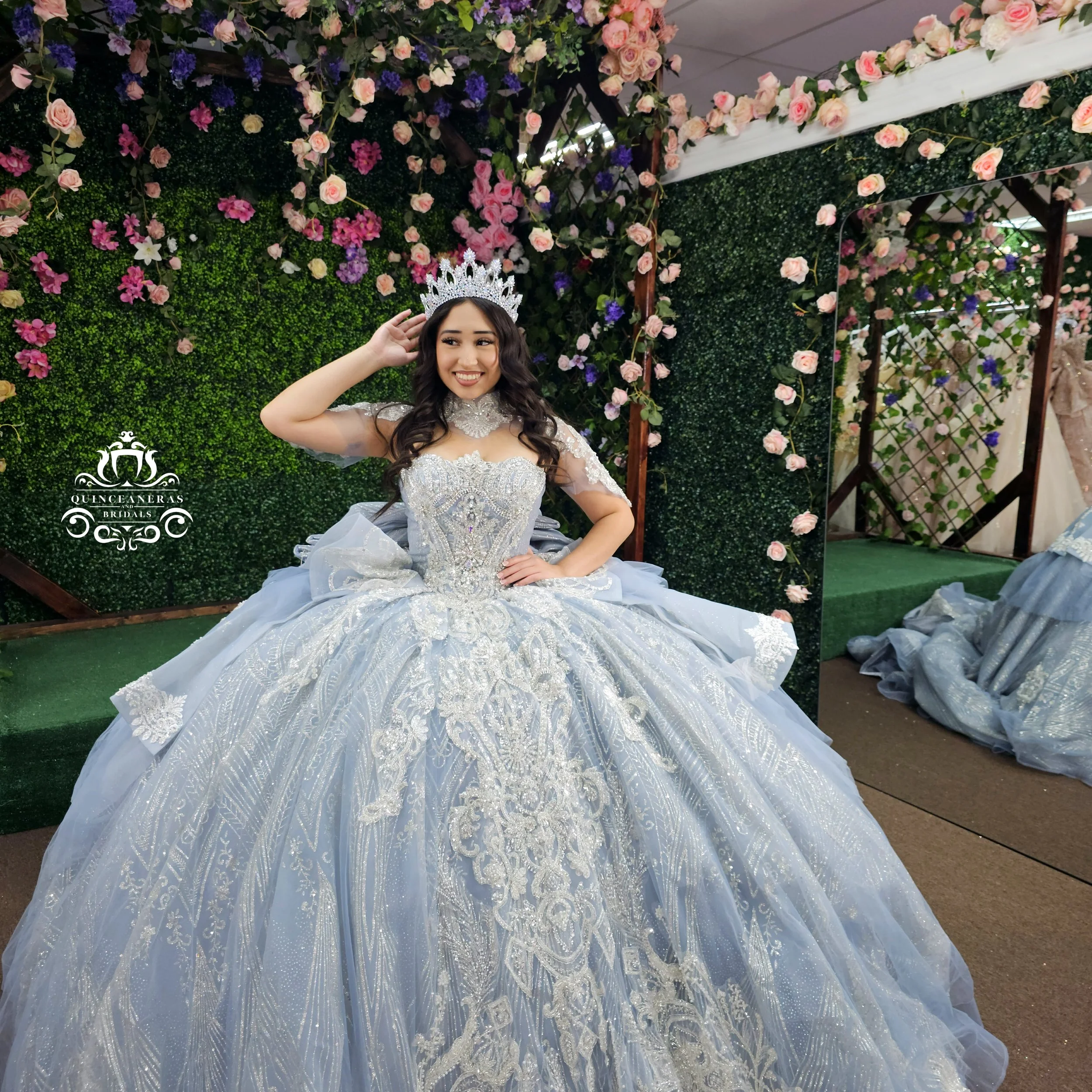 A woman in a silver and white ornate ball gown, wearing a crown, smiling and touching her hair, surrounded by pink and purple flowers on greenery backdrop.