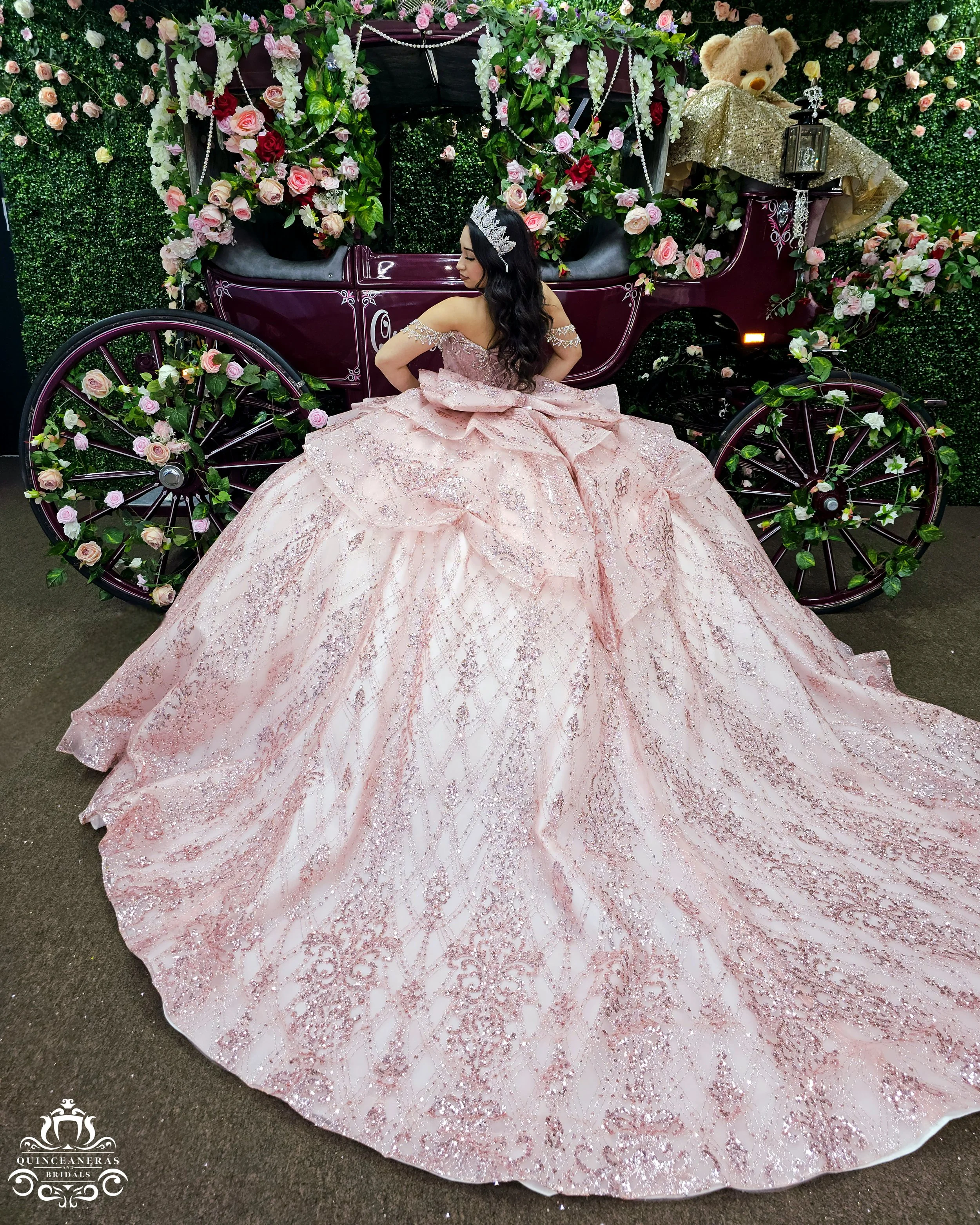 A woman in a pink, embellished quince dress with a full skirt and off-shoulder design, standing in front of a carriage decorated with pink and red roses, greenery, pearls, and a teddy bear with a gold dress in the background.