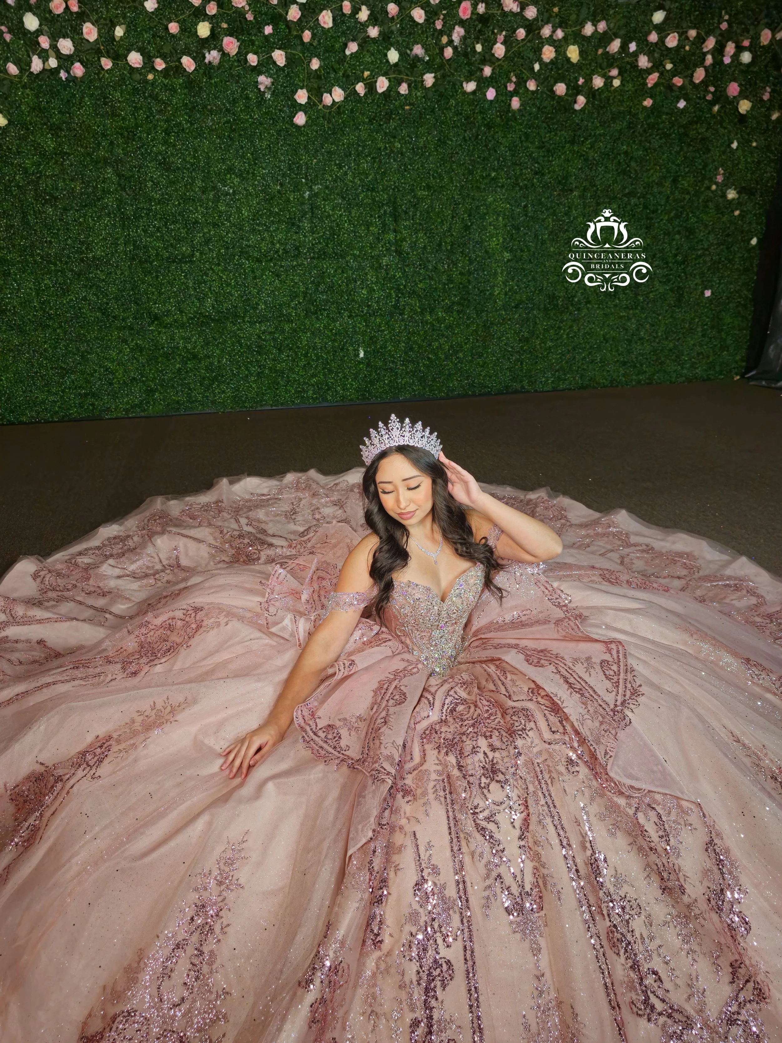 Young woman dressed in an elaborate pink quince dress with intricate beadwork and embroidery, wearing a tiara and jewelry, sitting on the floor against a green hedge backdrop, with the logo 'Quinceañeras and Bridals' visible on the wall.