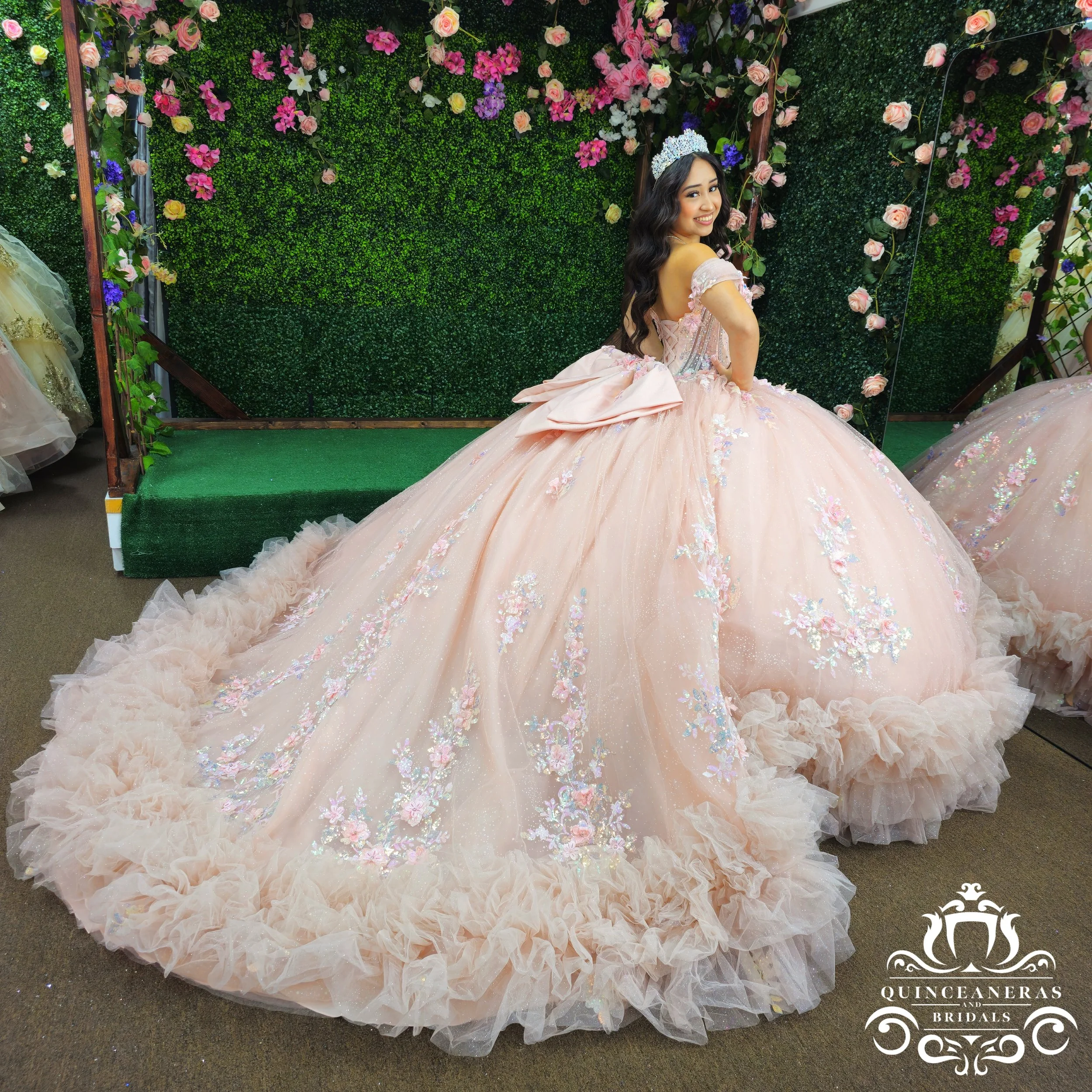 A woman in a pink quince dress with floral embroidery and a tiara, sitting on a pastel-colored, ruffled tulle dress in front of a green floral backdrop with pink and purple flowers.