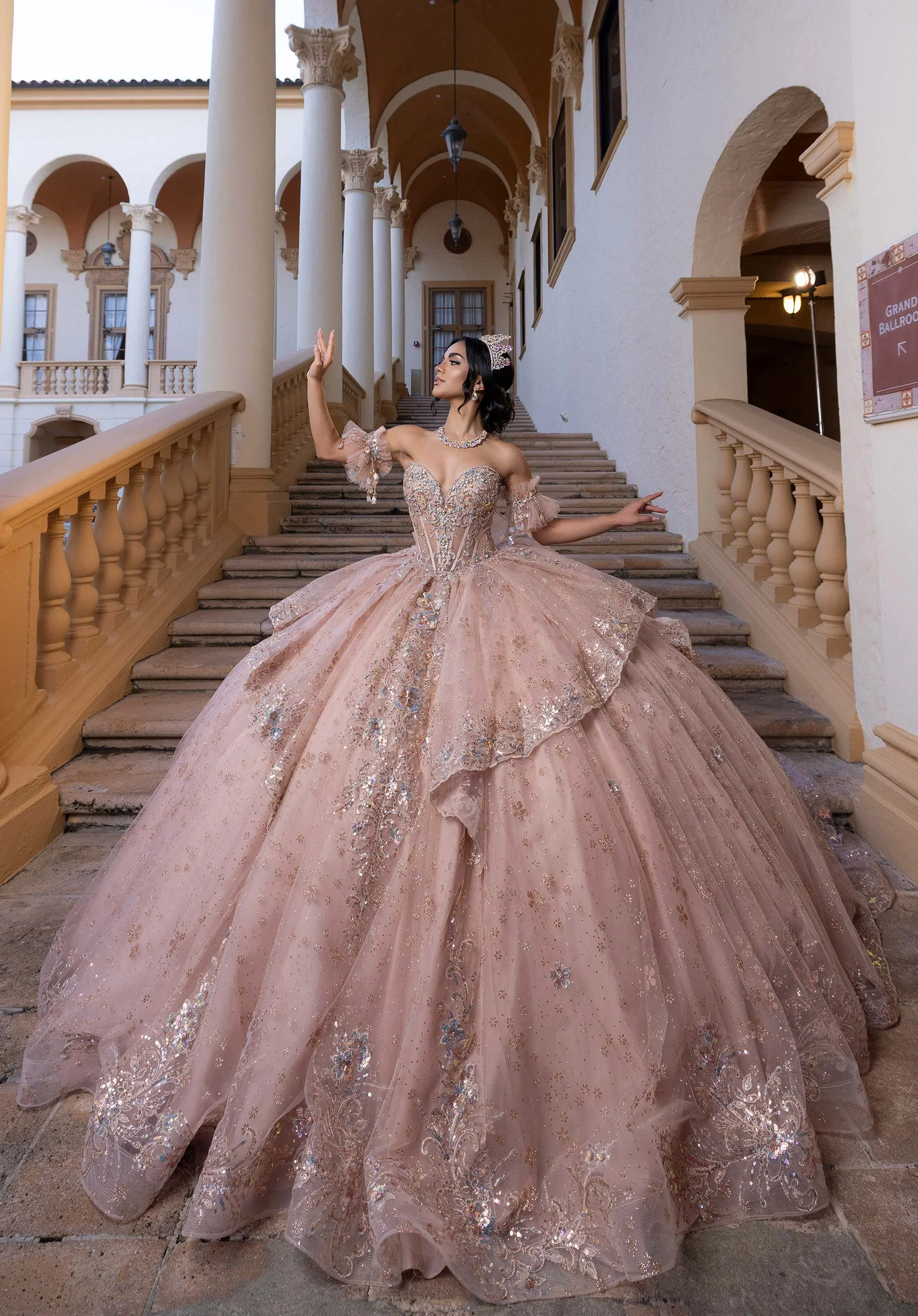 A woman wearing an elaborate pink quince dress with intricate embroidery, standing on a grand staircase in a historic building with columns and arches.
