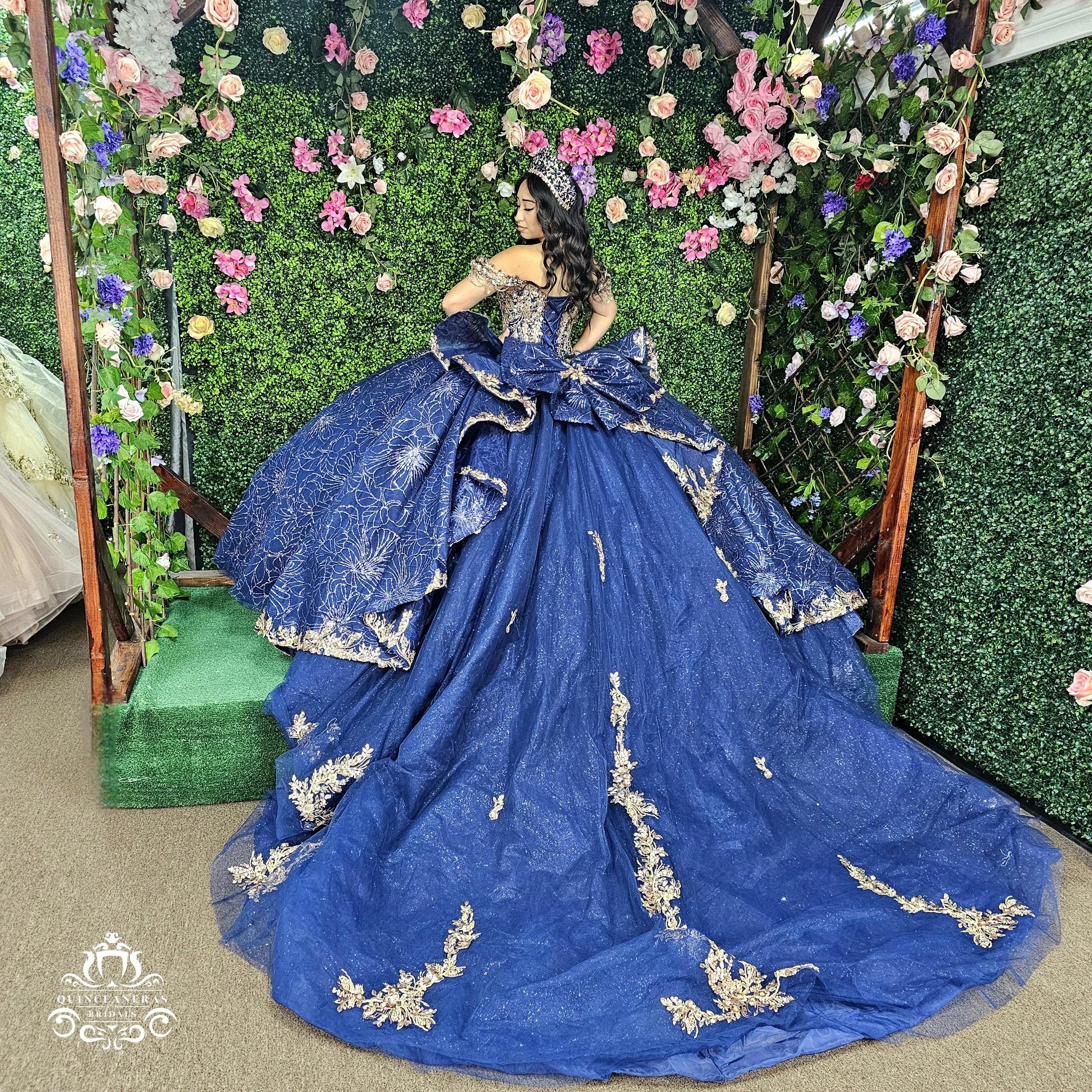 Young woman in a royal blue quinceañera dress with gold embroidery, standing in front of a green floral wall. The dress features a large bow and off-shoulder design, with matching headpiece.