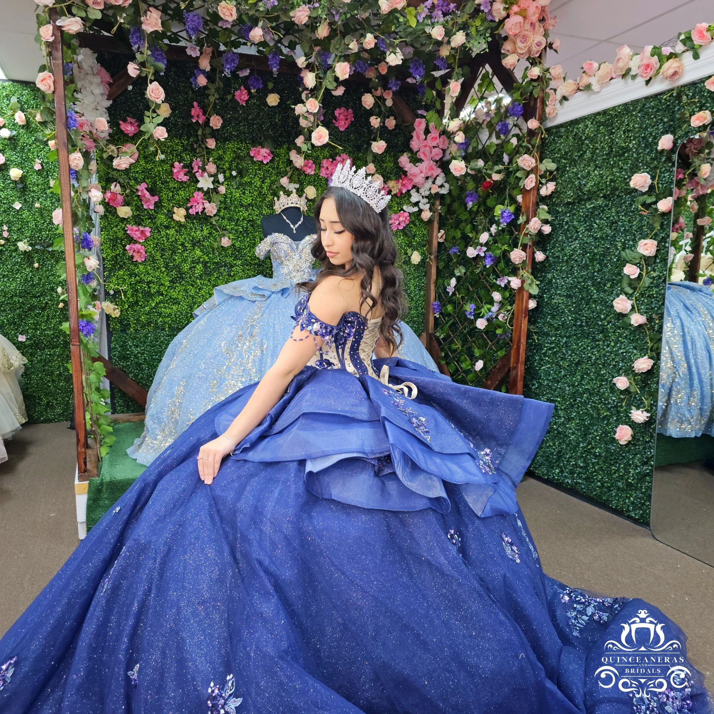 Young woman wearing a royal blue quince ball gown and a tiara sitting in front of a display of pastel pink and purple flowers and greenery with a mirror showing her reflection.